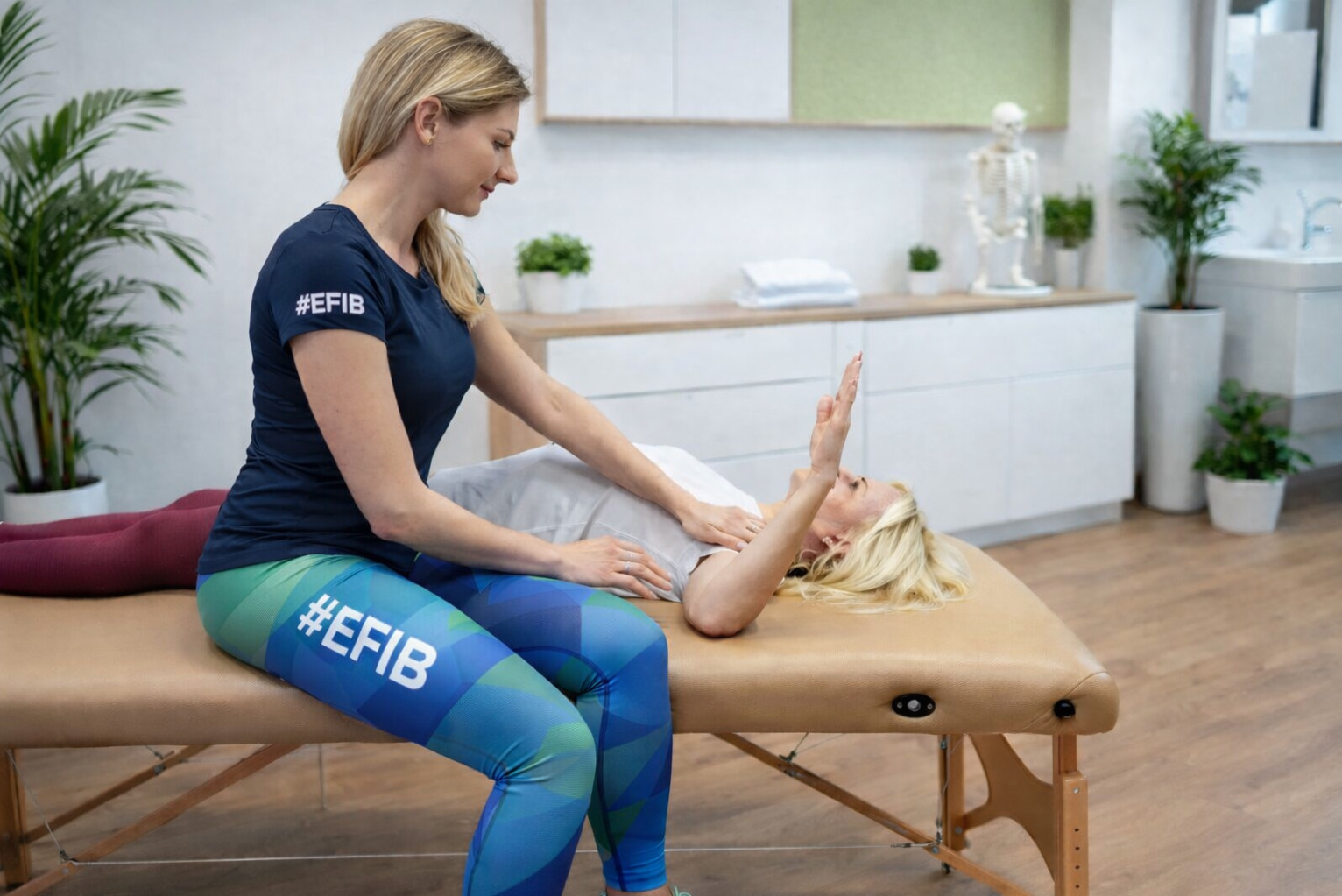 Kobieta w sportowym stroju wykonuje masaż klatki piersiowej pacjentki leżącej na kozetce w jasnym gabinecie z roślinami i modelem szkieletu.