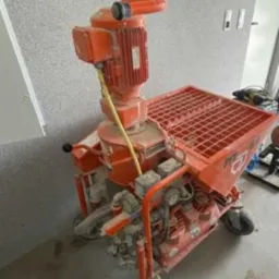 Pomarańczowa, używana maszyna do tynkowania PFT G4 z widocznymi śladami zaprawy i kurzu, stojąca na szarej podłodze w pomieszczeniu w trakcie prac wykończeniowych.