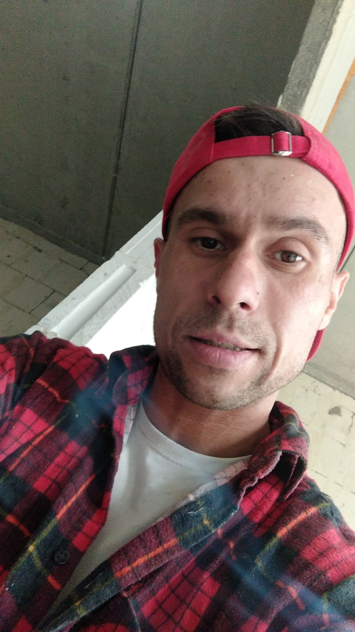 Autoportret pracownika budowlanego z Chrzanowa w czerwonej flanelowej koszuli i czerwonej czapce z daszkiem, z widocznym surowym betonowym sufitem w tle.