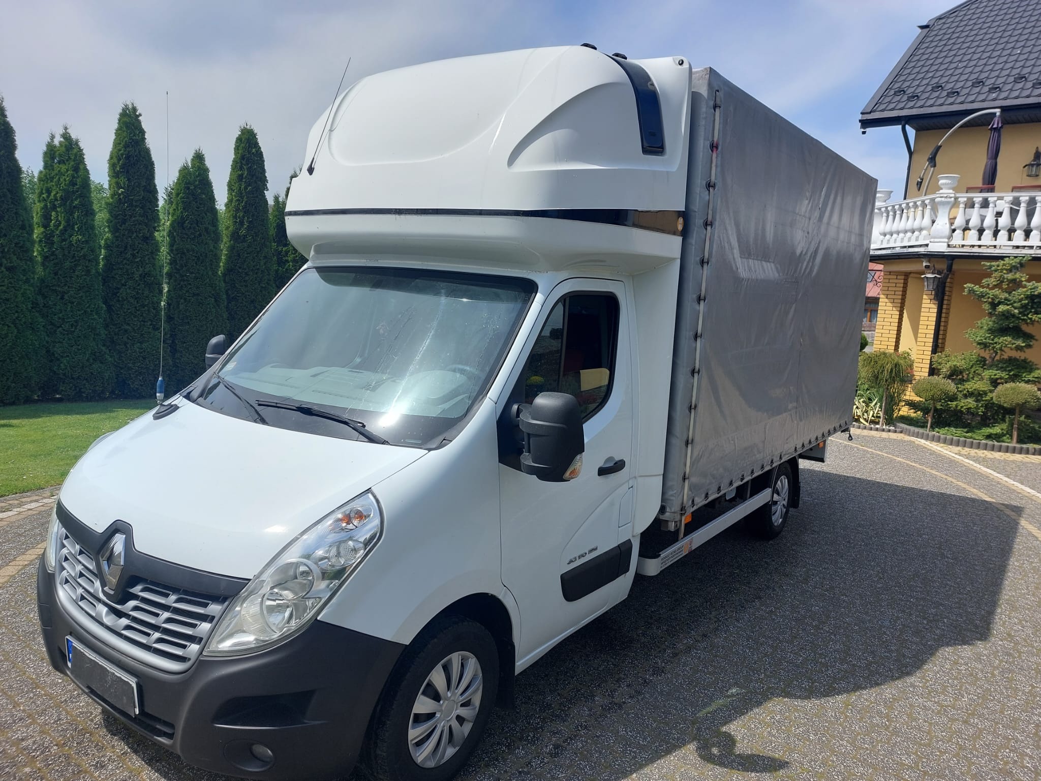Biały samochód dostawczy Renault Master z plandeką na tle zielonych tuj i żółtego domu z białymi balustradami.