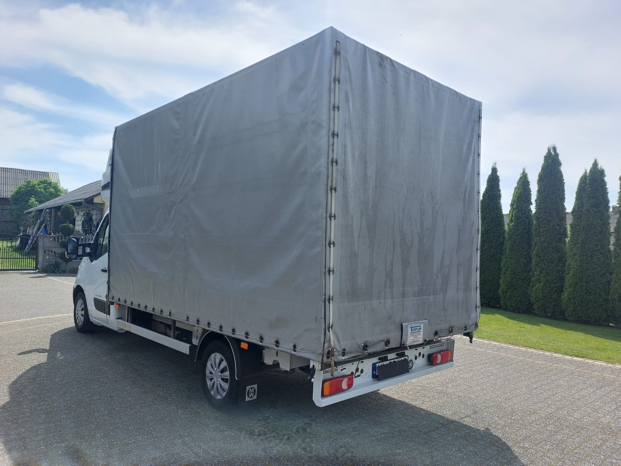 Biały samochód dostawczy z plandeką, widok z tyłu na parkingu, z widocznymi elementami mocowania plandeki i logo producenta pojazdu.