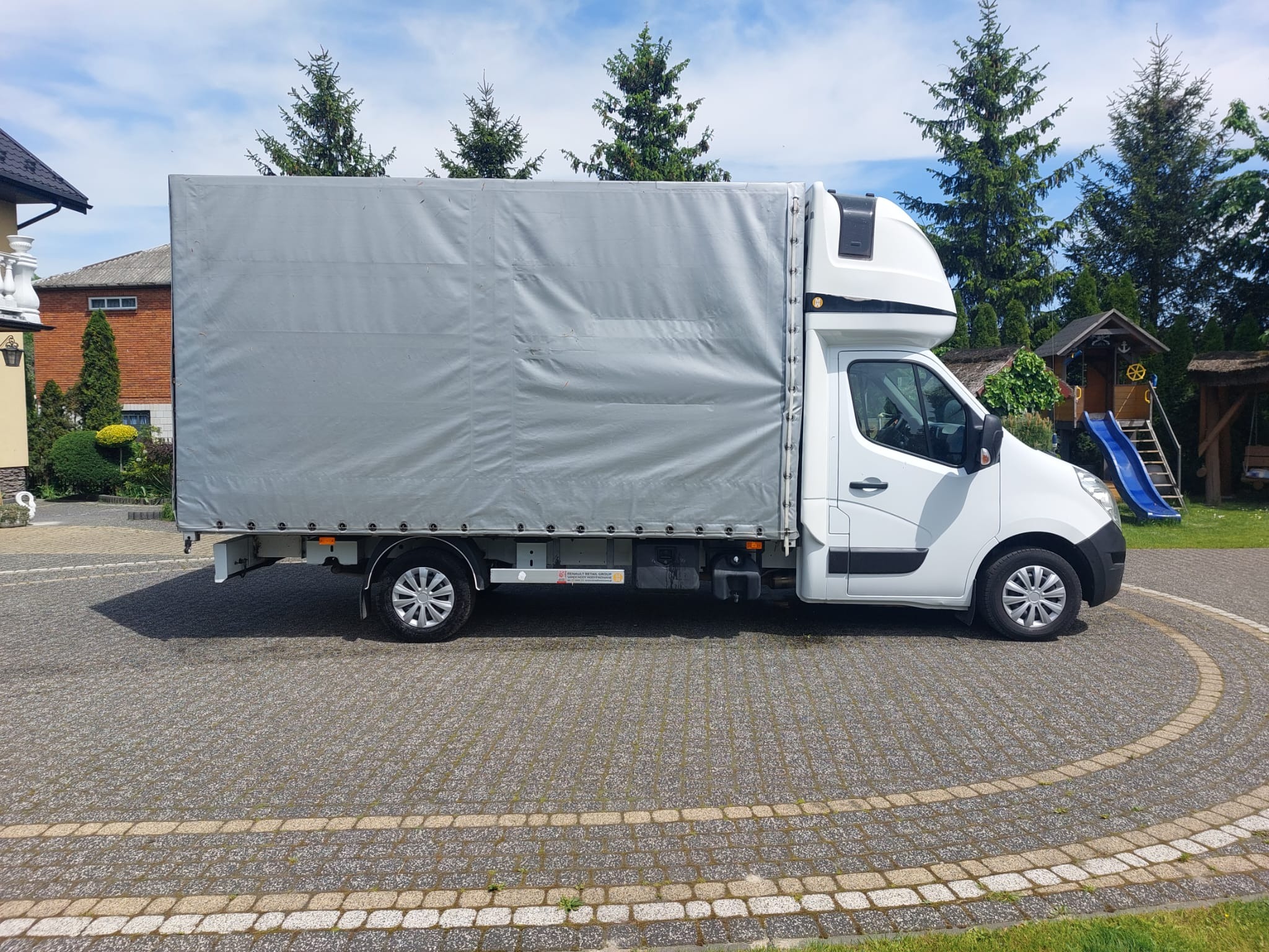 Biały samochód dostawczy z plandeką zaparkowany na kostce brukowej, w tle widoczne drzewa iglaste i plac zabaw.