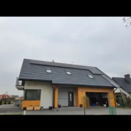 Dom jednorodzinny z zainstalowanymi panelami fotowoltaicznymi na dachu pokrytym szarą dachówką, widoczny również drabina oparta o dach.