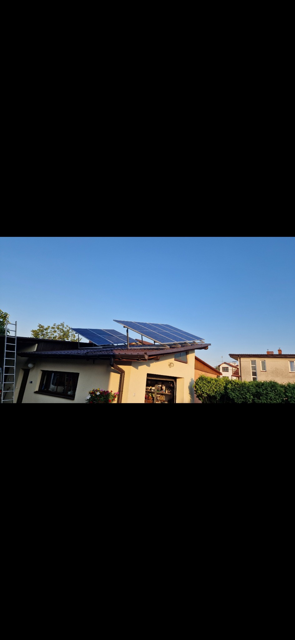 Panele słoneczne zamontowane na dachu garażu z drabiną opartą o ścianę, widok z dołu w słoneczny dzień.