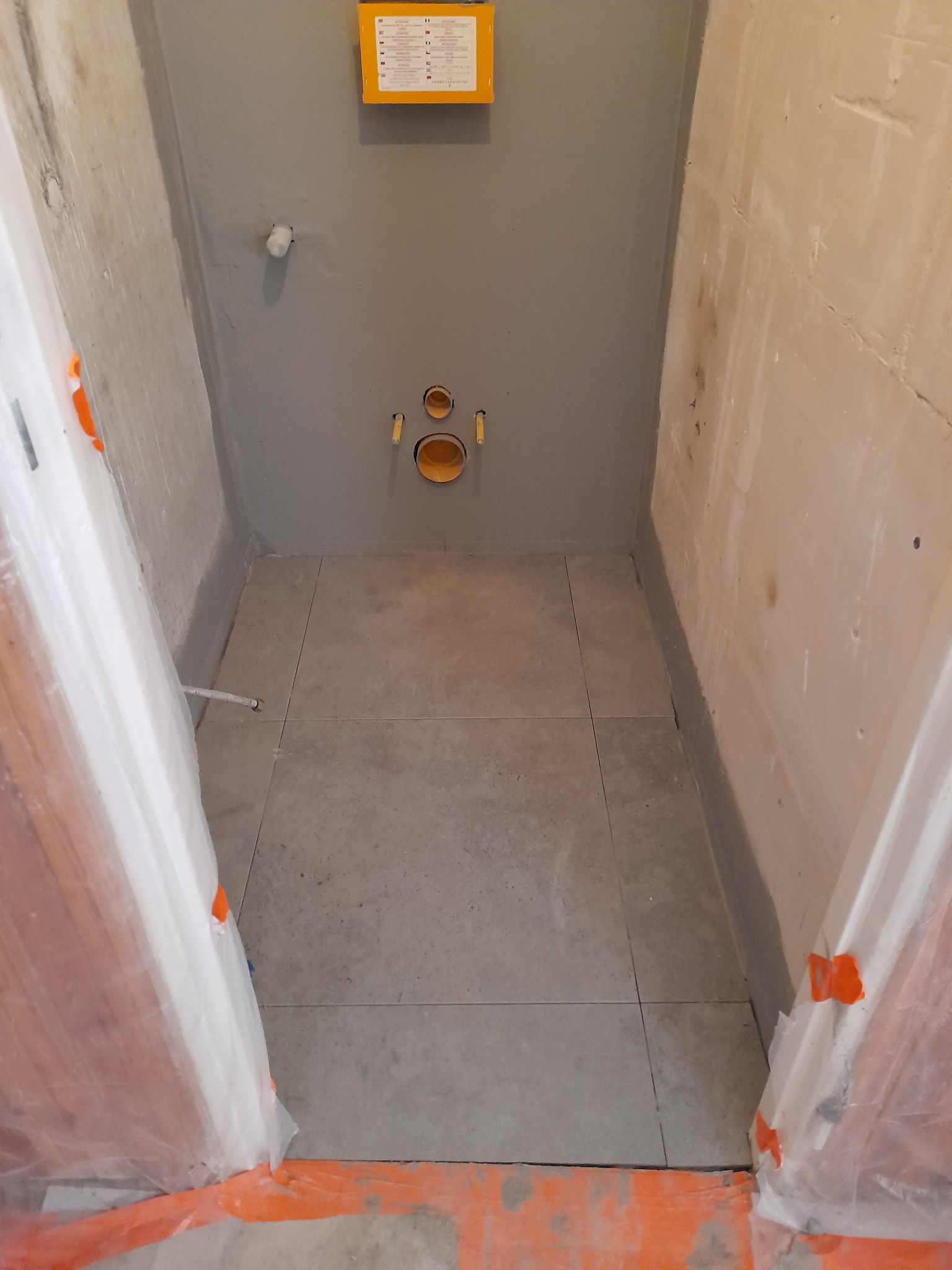 Przygotowane do montażu WC: szare płytki podłogowe w małej wnęce, zabezpieczone folią drzwi, widoczne przyłącza wodno-kanalizacyjne, szara ściana z żółtą skrzynką instalacyjną.