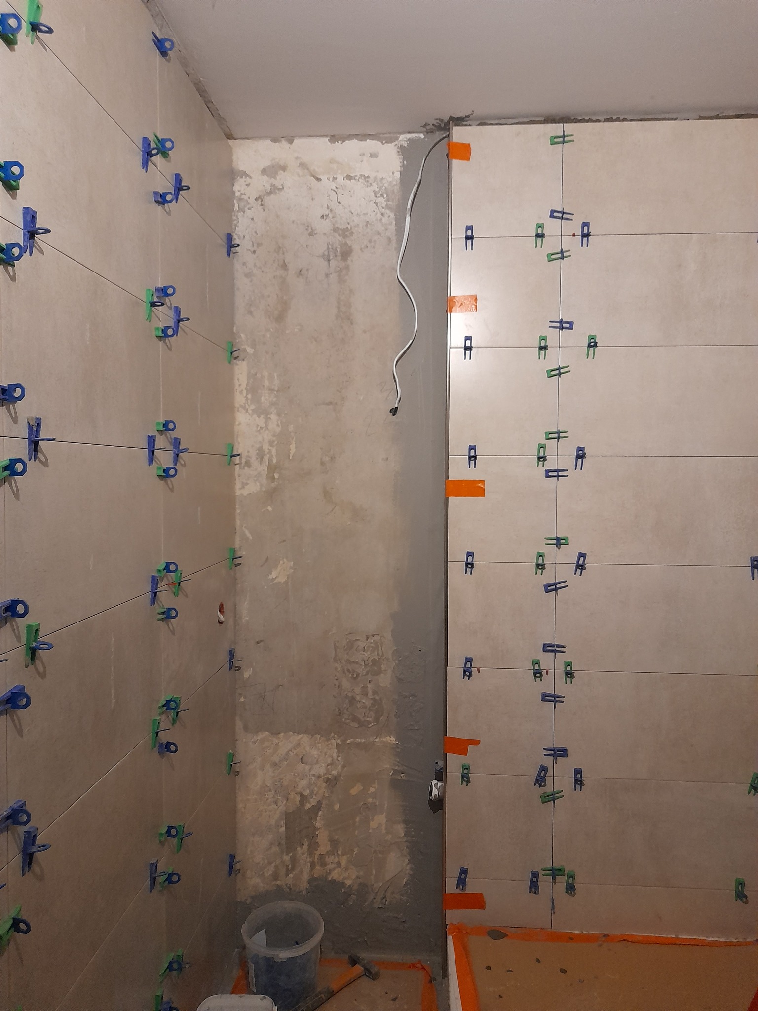 Ściana łazienki w trakcie kładzenia jasnych płytek, z użyciem systemu poziomowania płytek w kolorach niebieskim i zielonym oraz sznurków do wyznaczania linii, widoczny surowy fragment ściany obok.