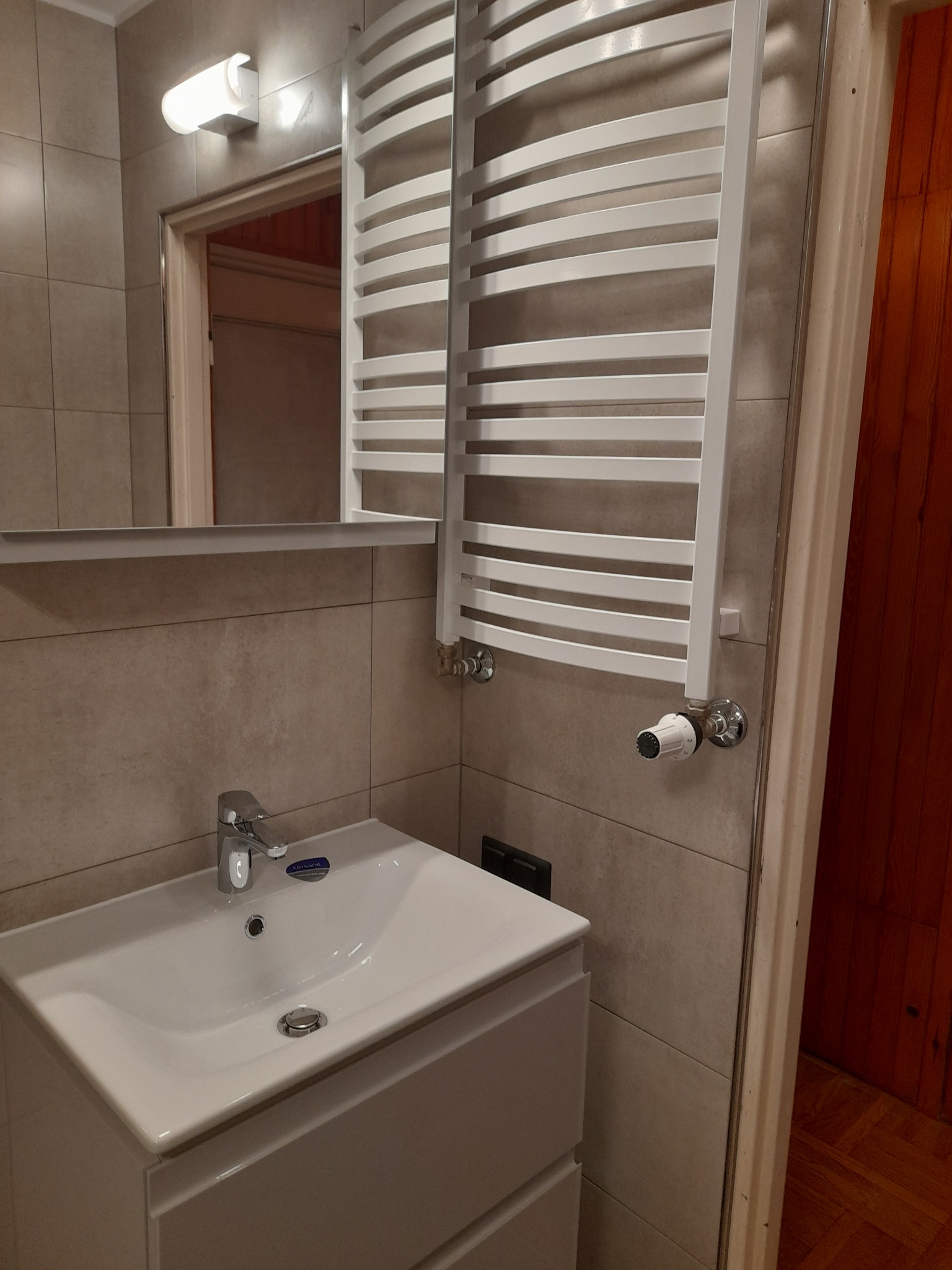 Biała umywalka z szafką, chromowana bateria i grzejnik drabinkowy w łazience wyłożonej beżowymi płytkami, widoczne odbicie w lustrze.