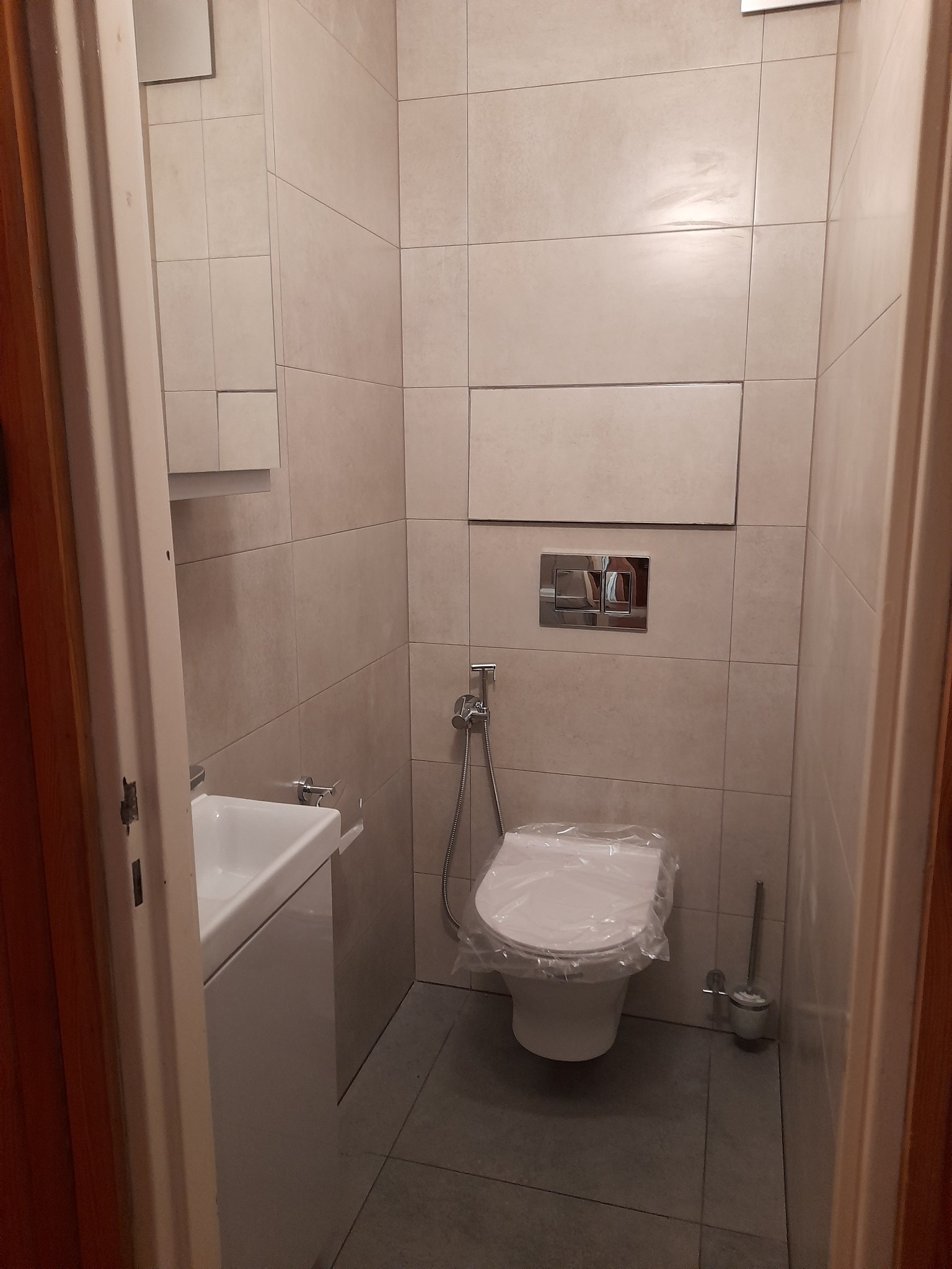 Wąska toaleta z beżowymi płytkami, umywalką z szafką i podwieszaną miską WC zabezpieczoną folią ochronną, widok zza otwartych drzwi.