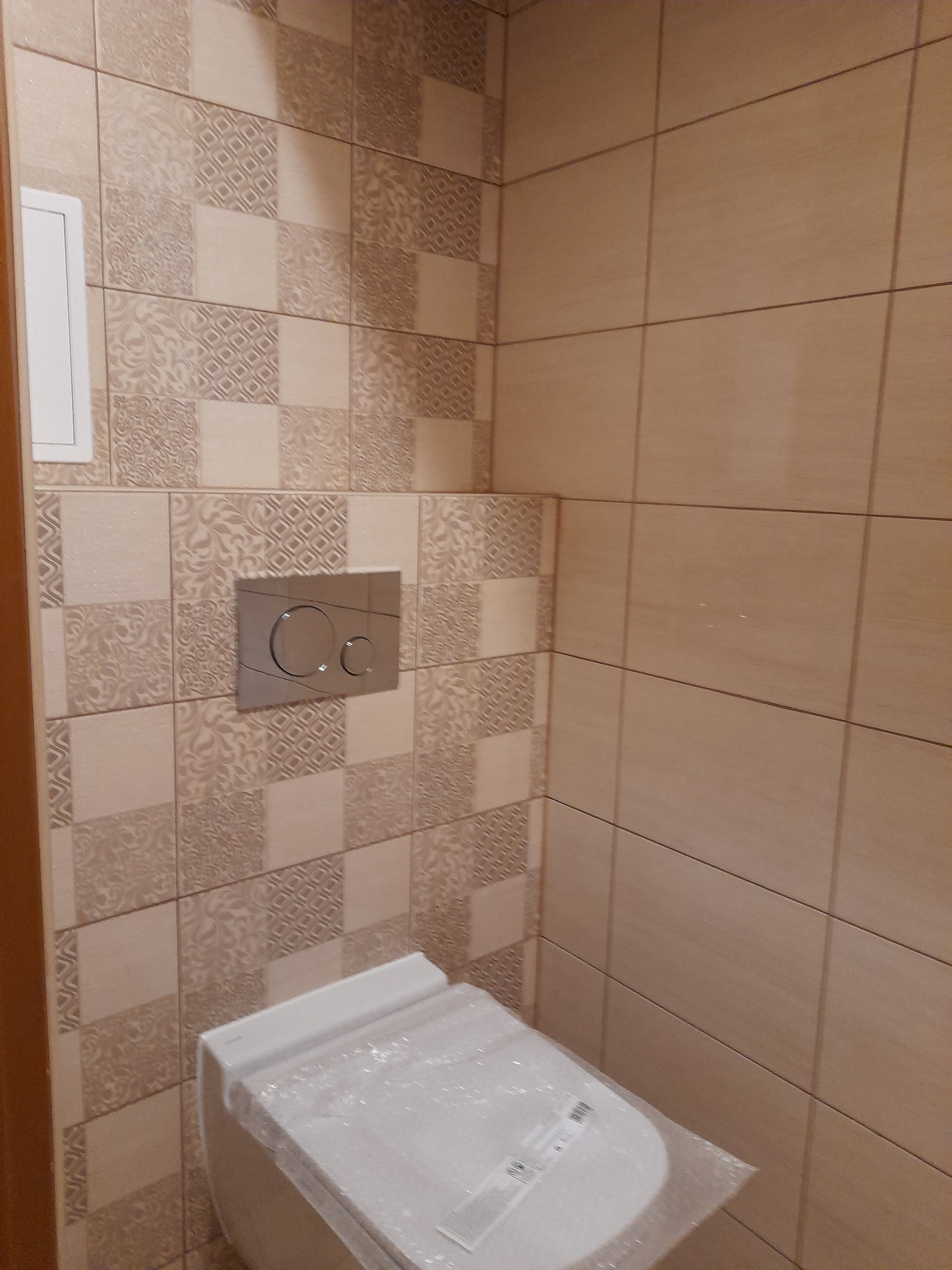 Nowa, biała toaleta, jeszcze w folii ochronnej, zamontowana w łazience z beżowymi płytkami o zróżnicowanym wzorze, widoczny metalowy przycisk spłuczki.