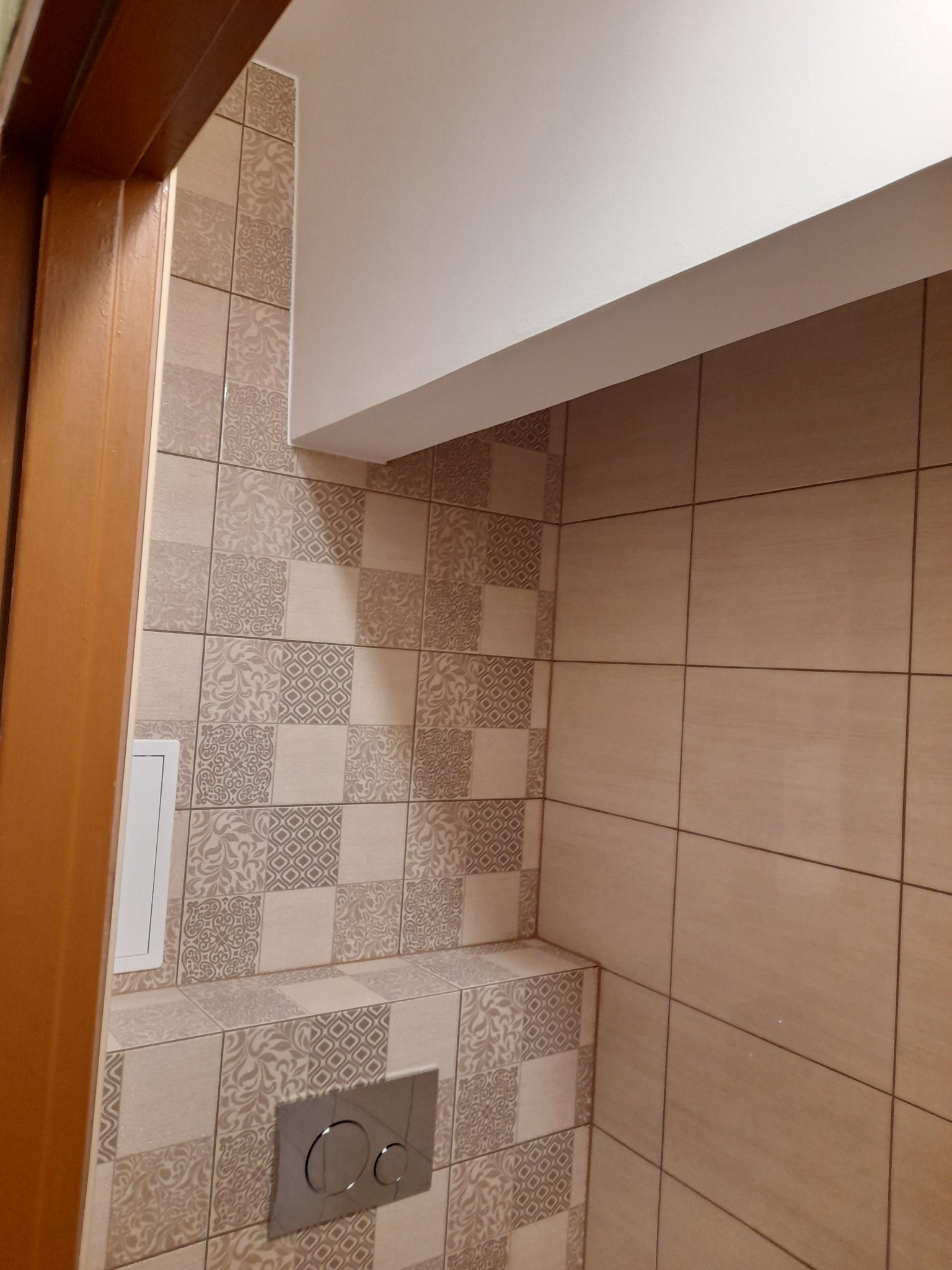 Wykończona łazienka z beżowymi płytkami w patchworkowy wzór, widoczny przycisk spłuczki toalety podwieszanej i fragment ościeżnicy drewnianej.