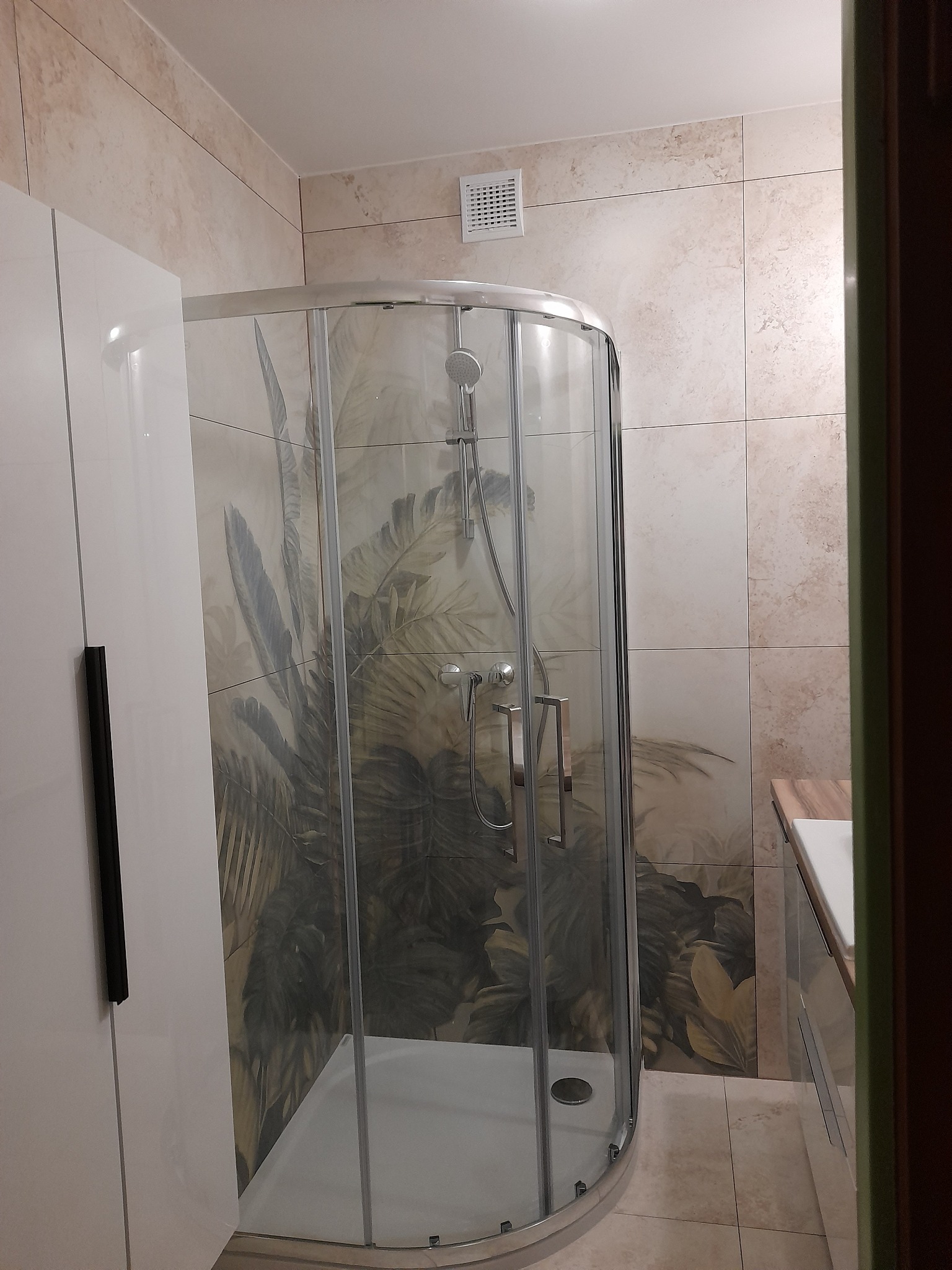 Zaokrąglona kabina prysznicowa z wzorem roślinnym na ścianach, obok biała szafka łazienkowa z czarnym uchwytem i beżowe płytki podłogowe.