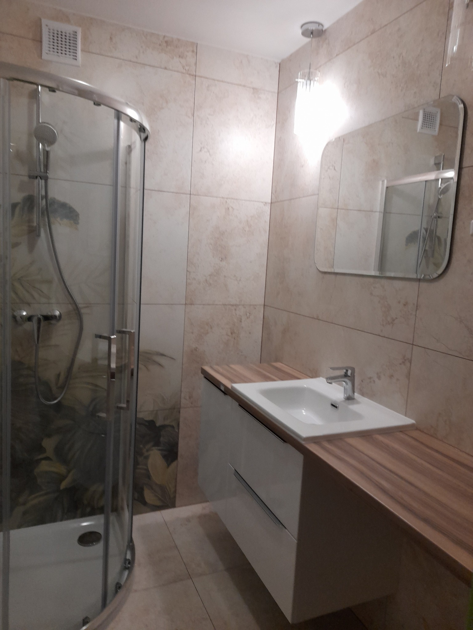 Nowoczesna łazienka z kabiną prysznicową z motywem roślinnym, umywalką nablatową z drewnianym blatem i lustrem z zaokrąglonymi rogami, wykończona beżowymi płytkami.