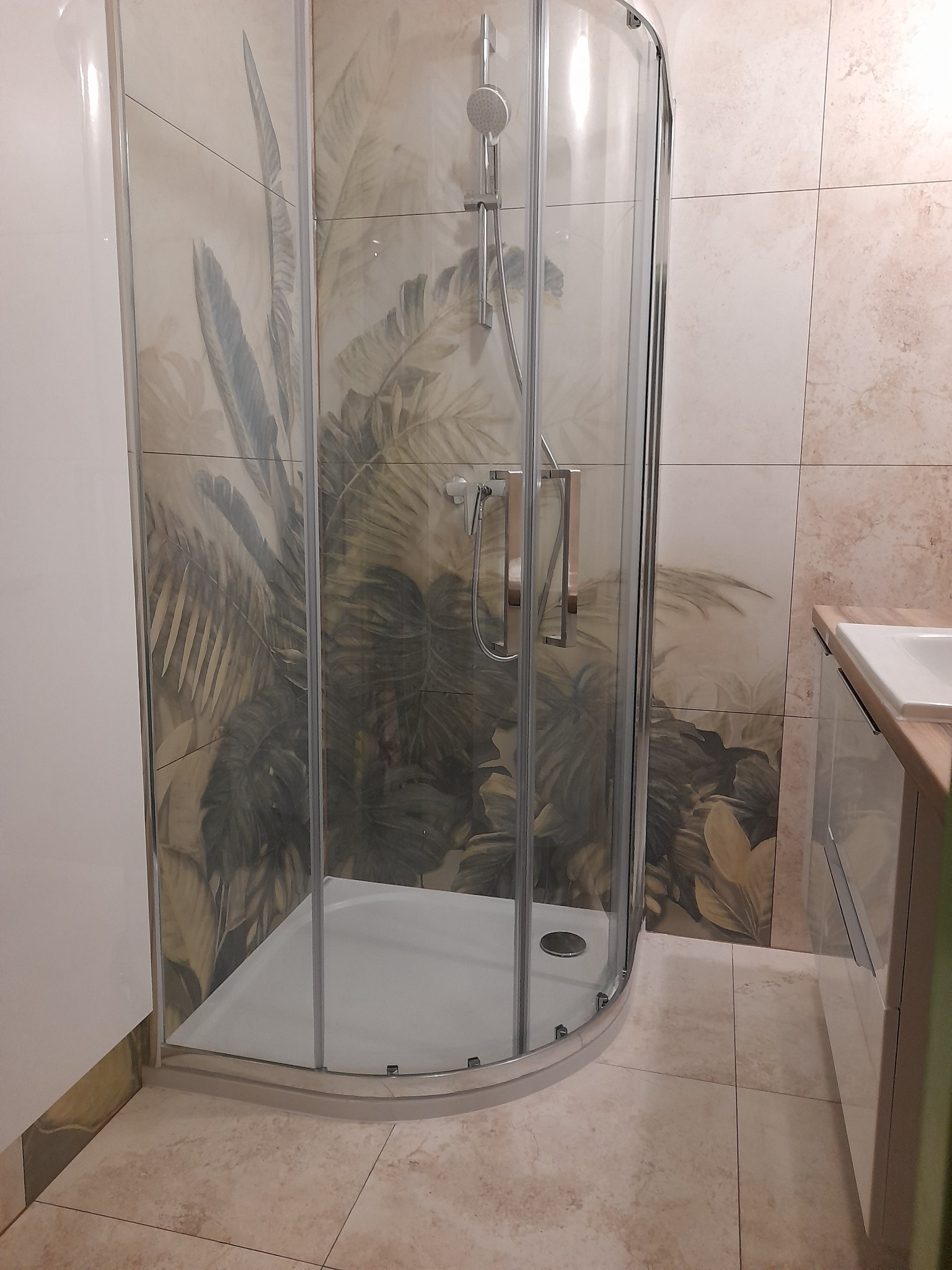 Kabina prysznicowa z motywem tropikalnych liści na płytkach w łazience w Warszawie, widoczna umywalka z szafką.