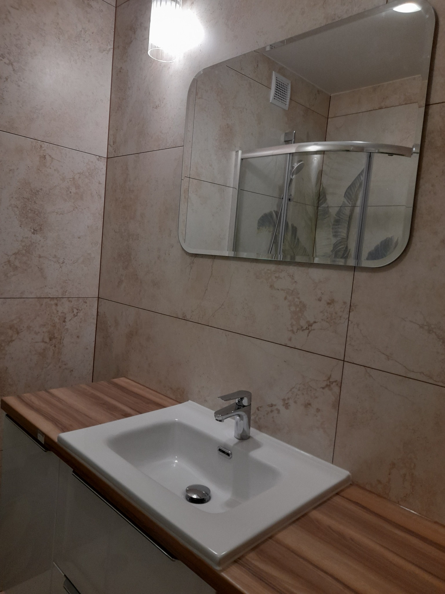 Umywalka ceramiczna z chromowaną baterią, osadzona na blacie imitującym drewno, na tle beżowych płytek imitujących marmur i lustra z widoczną kabiną prysznicową z motywem liści.