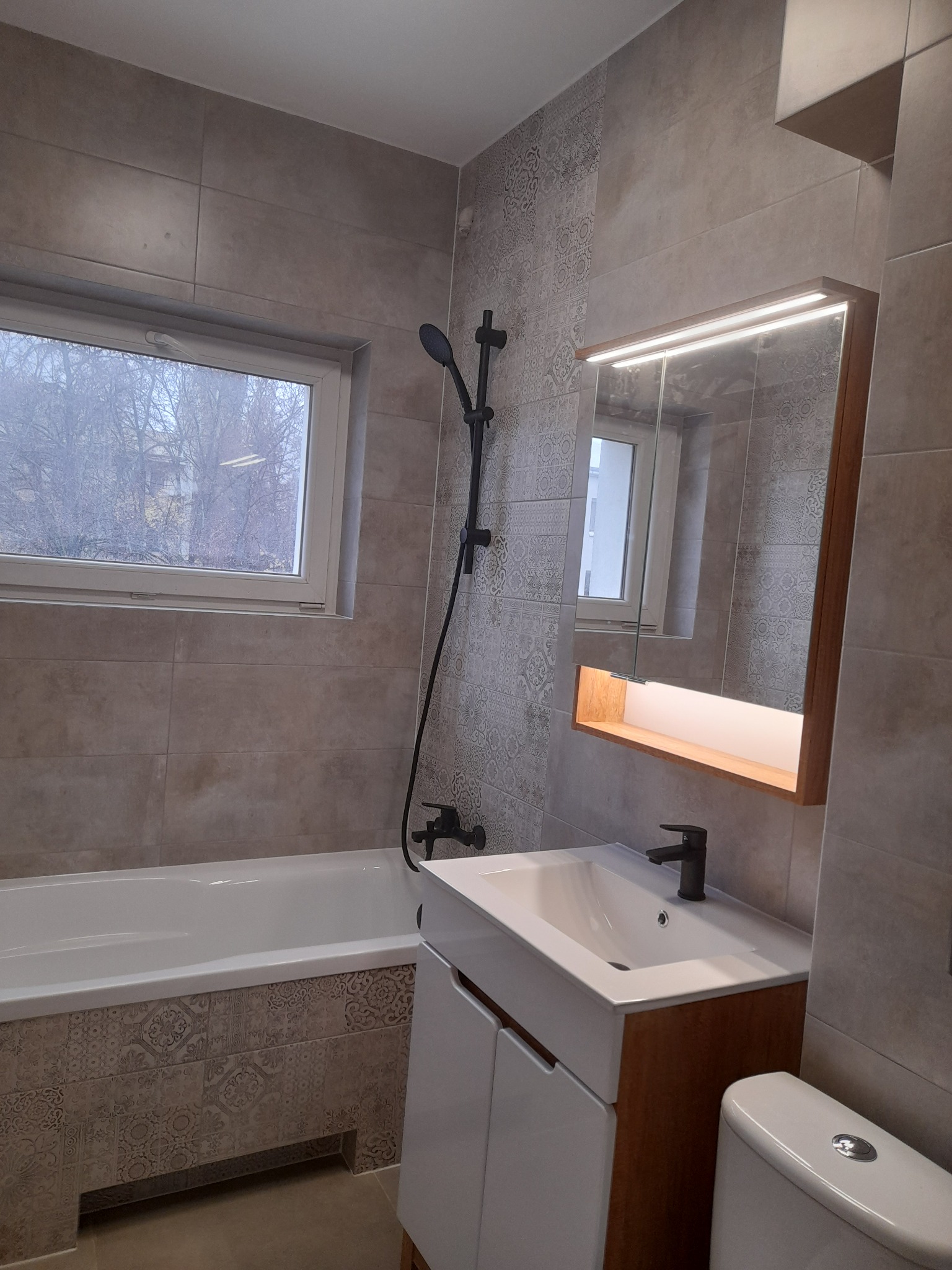 Nowoczesna łazienka z beżowymi płytkami, armaturą w kolorze czarnym, podświetlanym lustrem i oknem z widokiem na drzewa.