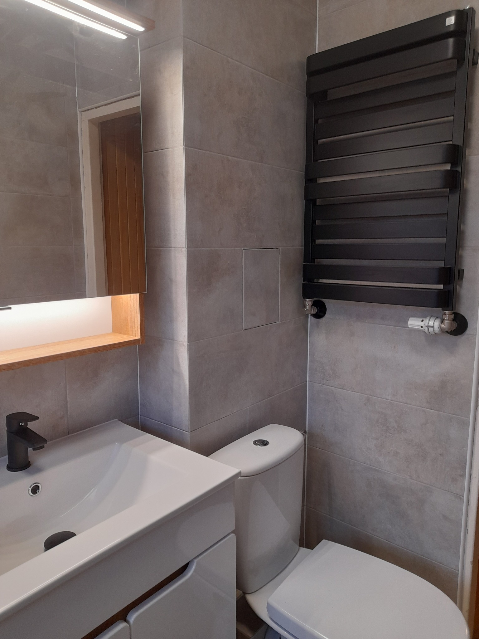 Nowoczesna łazienka z szarymi płytkami, białą umywalką z czarną armaturą, toaletą i grzejnikiem drabinkowym w kolorze czarnym.
