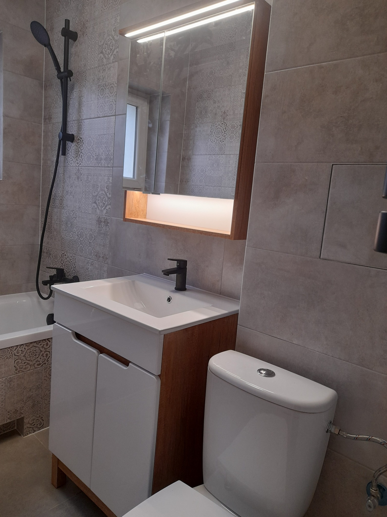 Nowoczesna łazienka z szarymi płytkami, białą umywalką z szafką, toaletą i czarną armaturą, oświetlone lustro nad umywalką.