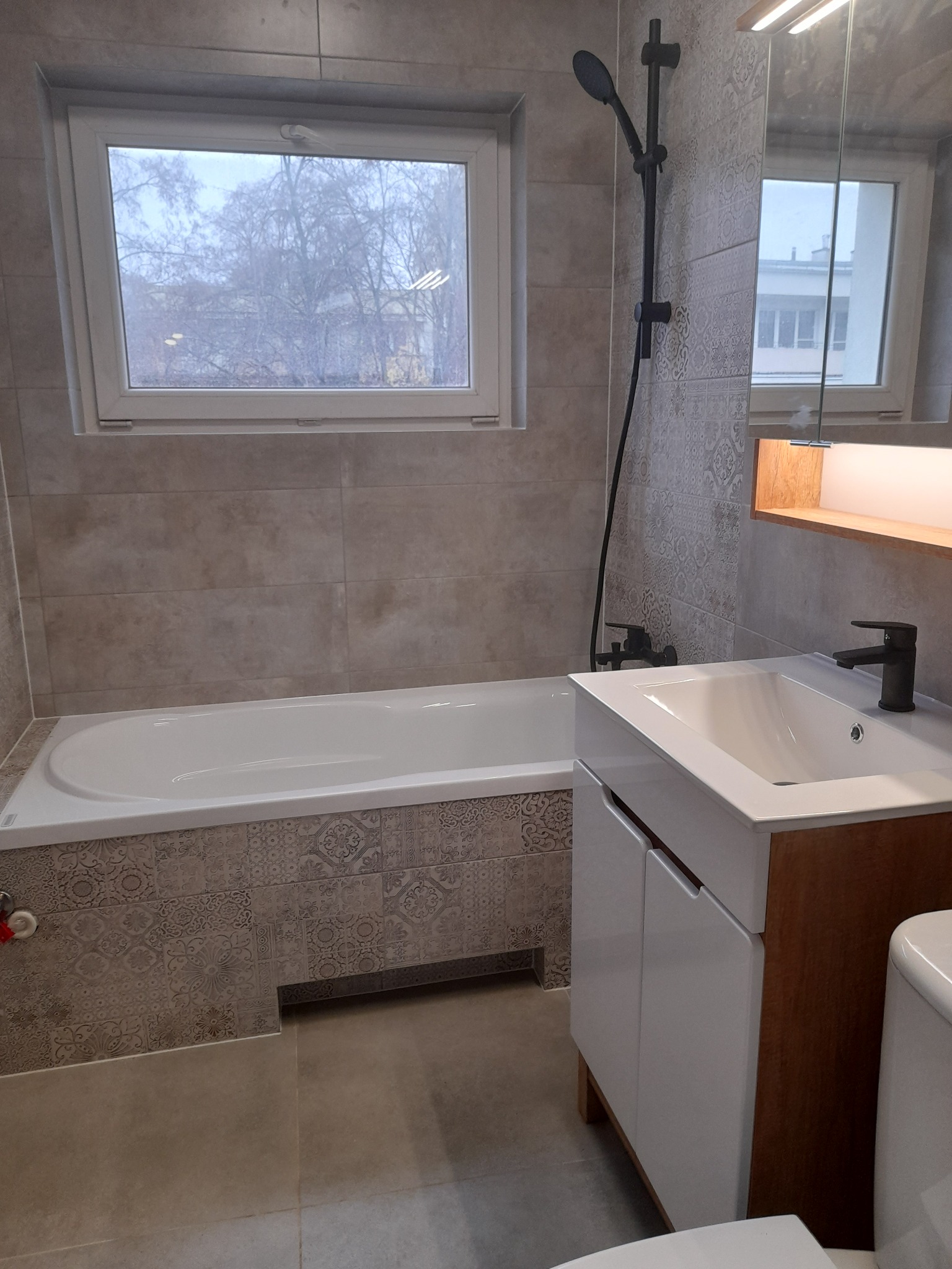 Nowoczesna łazienka z beżowymi płytkami, wanną obudowaną dekoracyjnymi kaflami, białą szafką pod umywalką z drewnianymi bokami oraz czarną armaturą, oświetlona ledowym światłem zza lustra.