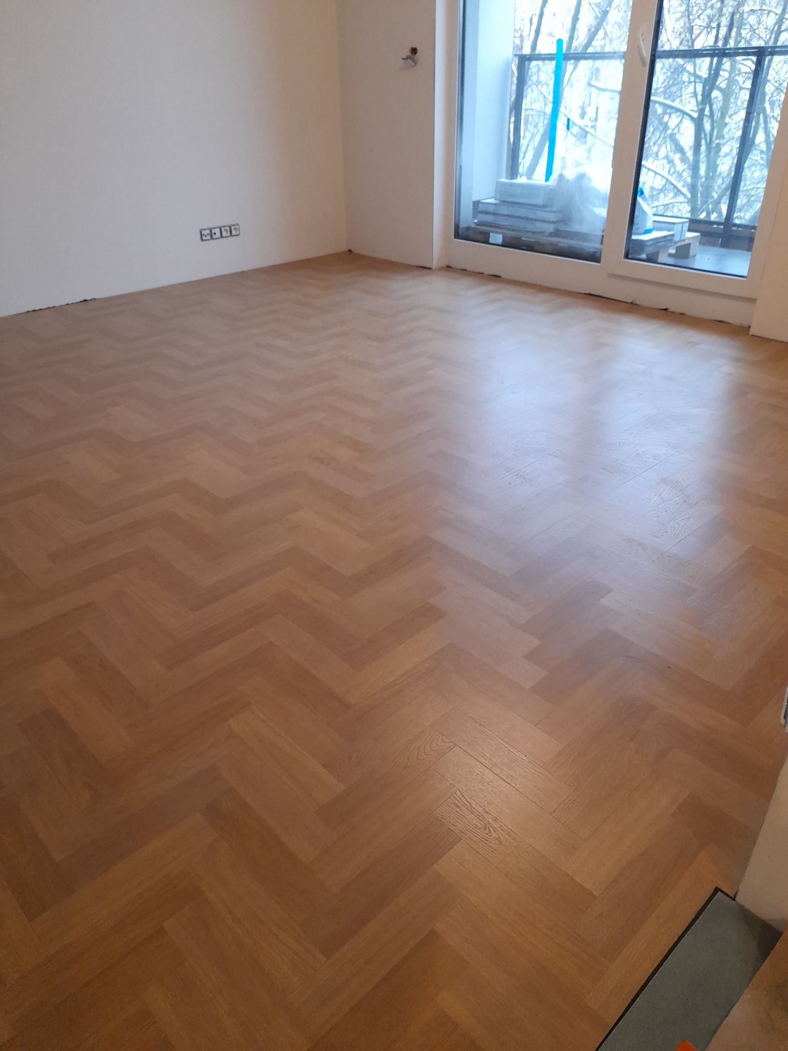 Nowo ułożony parkiet w jodełkę w pustym pomieszczeniu z dużym oknem, przez które widać ośnieżone drzewa.