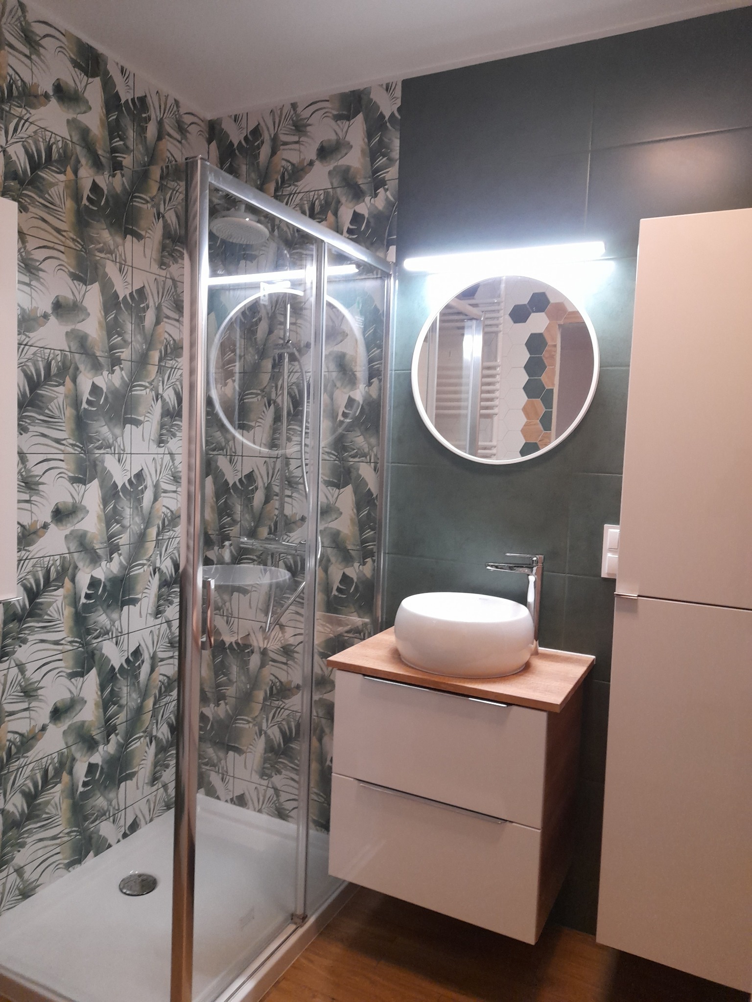 Nowoczesna łazienka z kabiną prysznicową, umywalką nablatową na szafce z drewnianym blatem, okrągłym lustrem podświetlanym LED i płytkami z motywem liści na ścianie.