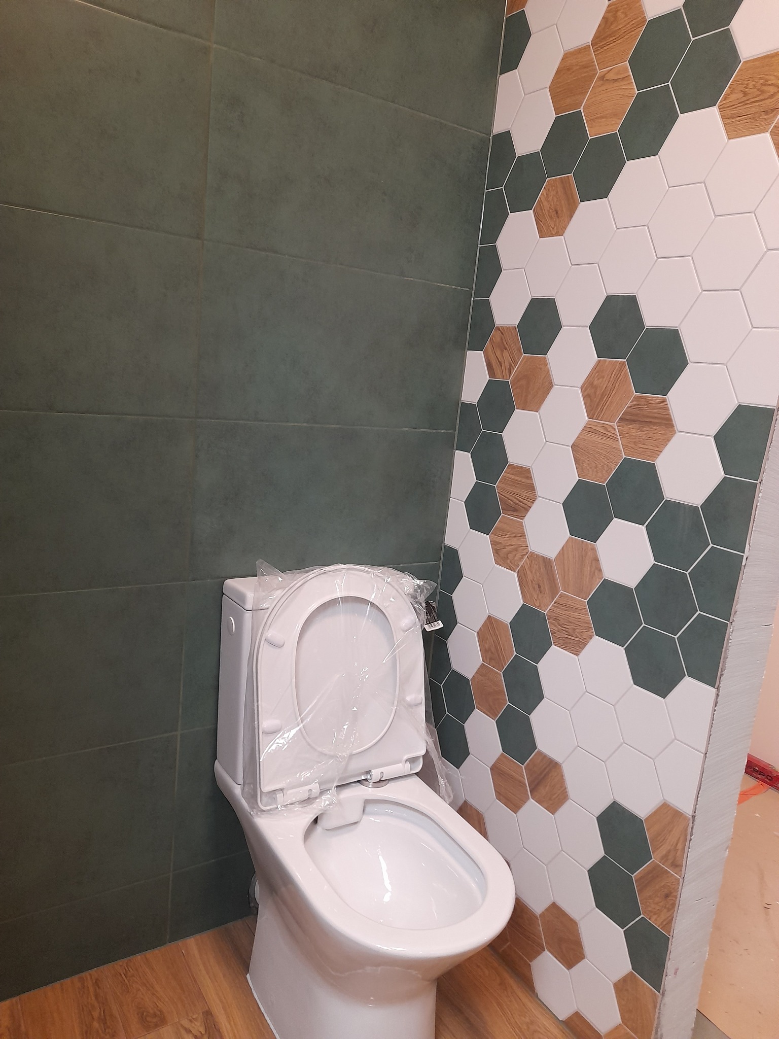 Nowa, biała toaleta w folii ochronnej na tle ściany z zielonych, brązowych i białych heksagonalnych płytek oraz ściany z ciemnozielonych płytek.