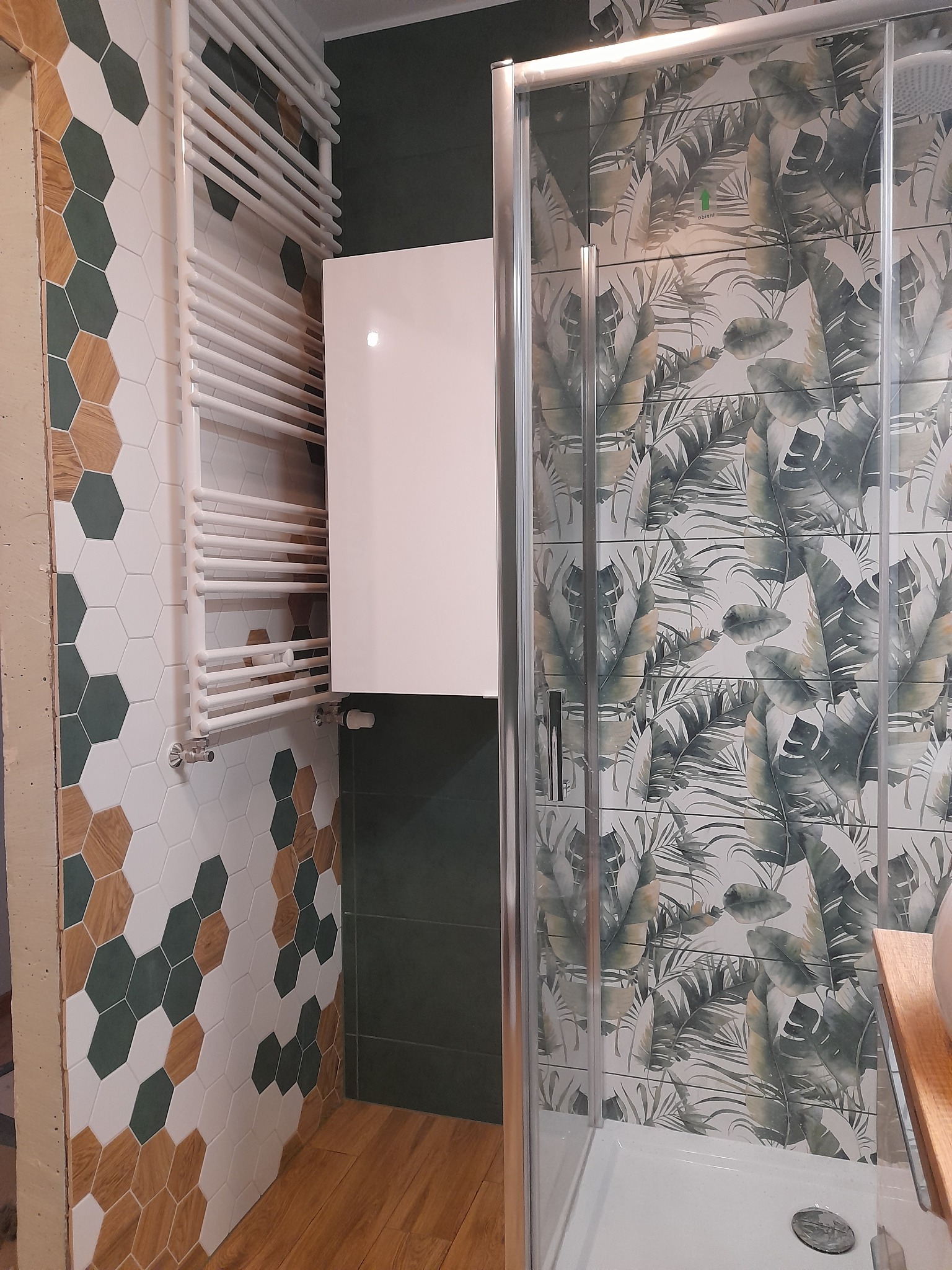 Nowoczesna łazienka w Warszawie z kabiną prysznicową z motywem liści, grzejnikiem drabinkowym na ścianie wyłożonej heksagonalnymi płytkami w odcieniach bieli, zieleni i drewna.