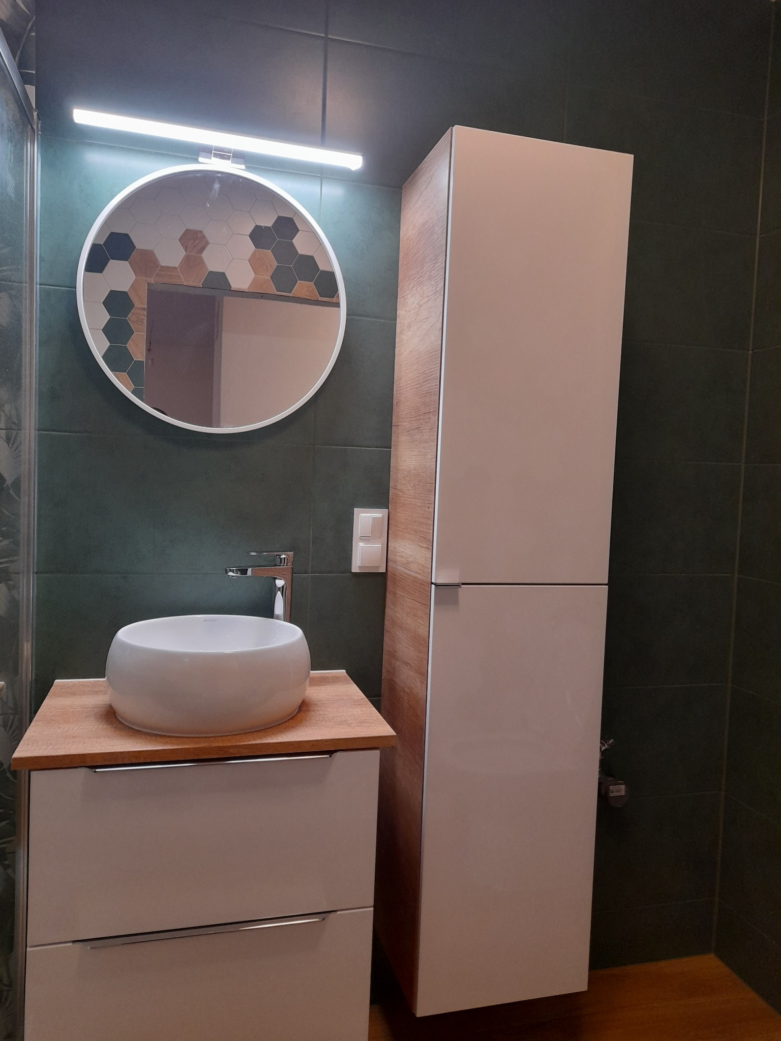 Nowoczesna łazienka z okrągłym lustrem z heksagonalnymi akcentami, umywalką nablatową i wysoką szafką w odcieniach bieli i drewna, na tle ciemnozielonych płytek.