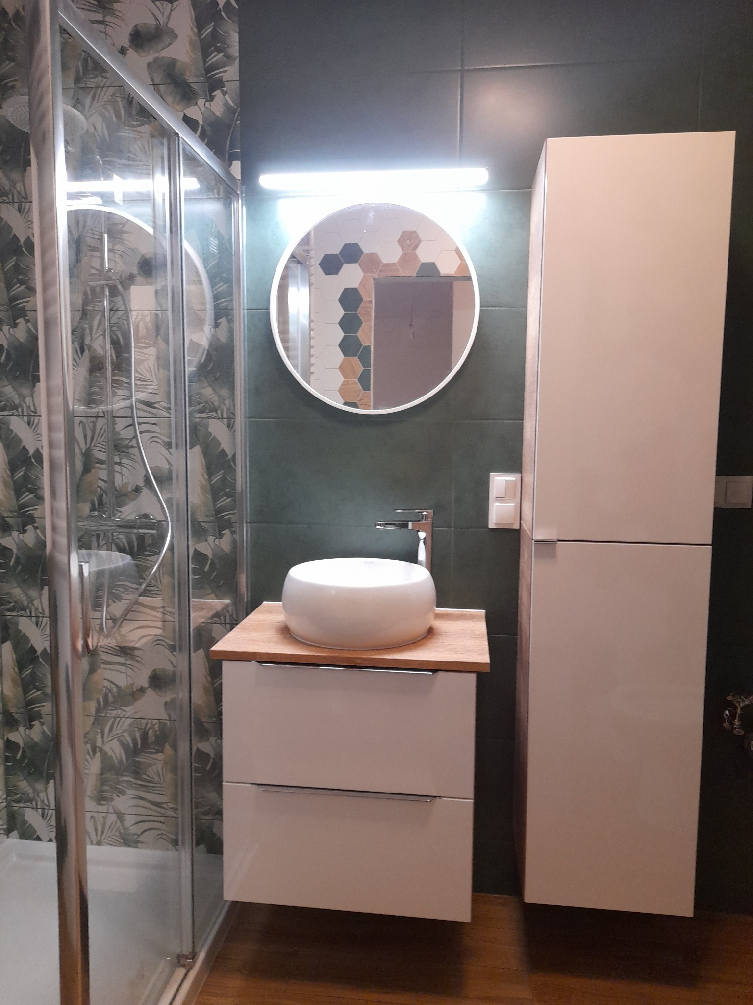 Nowoczesna łazienka z kabiną prysznicową z motywem liści, okrągłym lustrem podświetlanym LED i białą umywalką nablatową na szafce z drewnianym blatem.