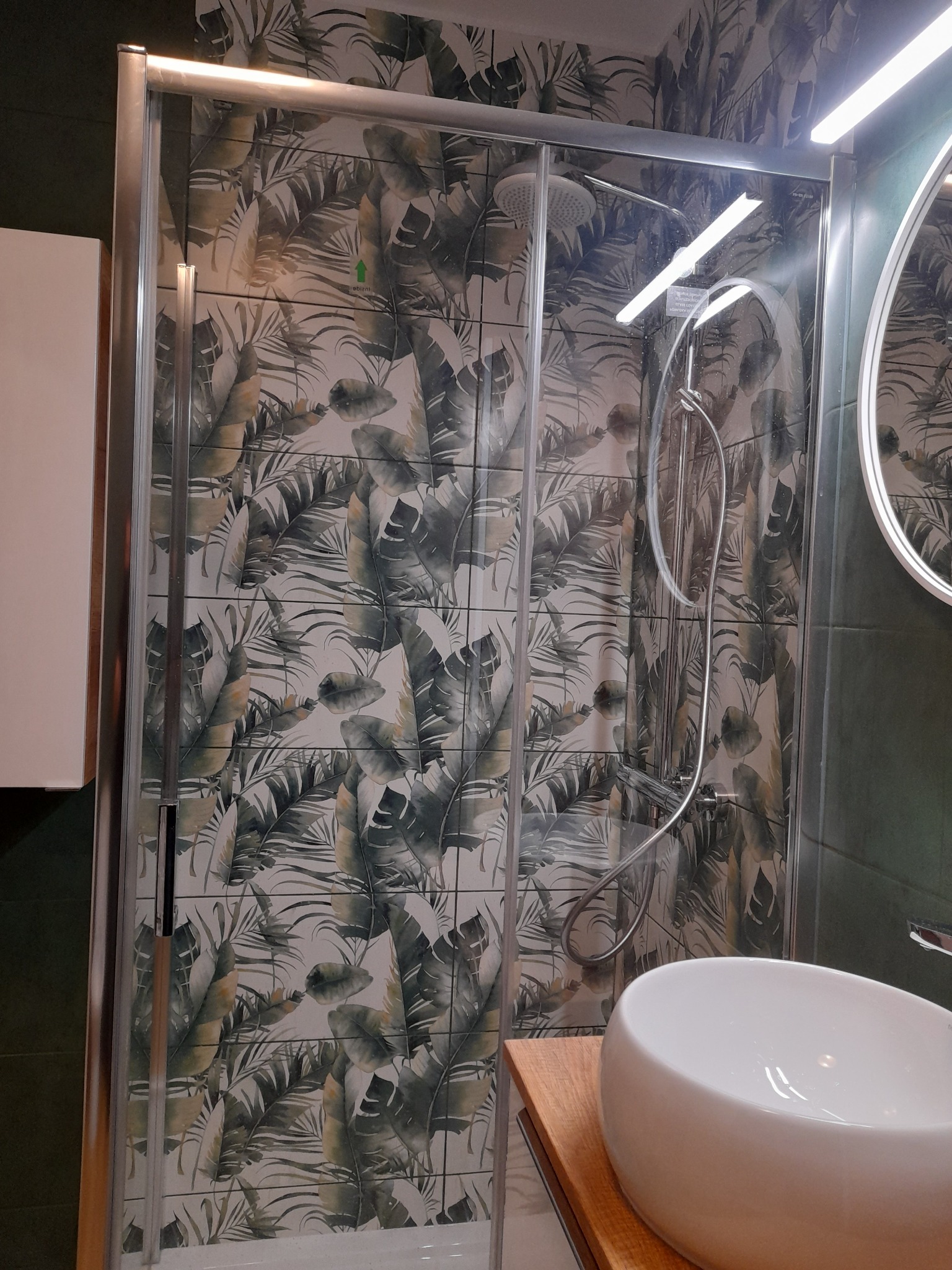 Kabina prysznicowa z przesuwnymi drzwiami na tle ściany wyłożonej płytkami z motywem liści palmowych, widoczna umywalka nablatowa i oświetlone lustro.
