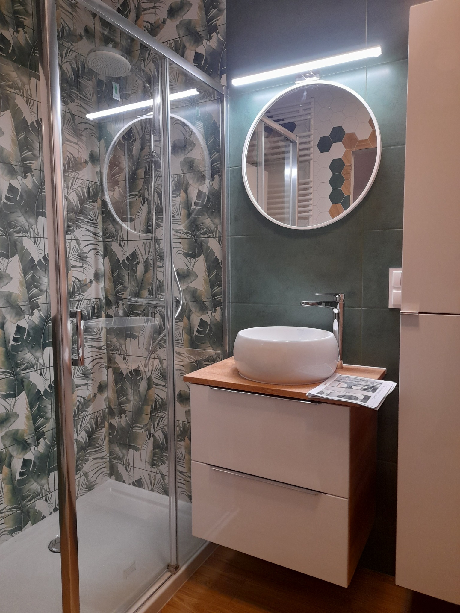 Nowoczesna łazienka po remoncie: kabina prysznicowa z deszczownicą, umywalka nablatowa na szafce z drewnianym blatem, okrągłe lustro z podświetleniem LED, płytki z motywem liści na ścianach.