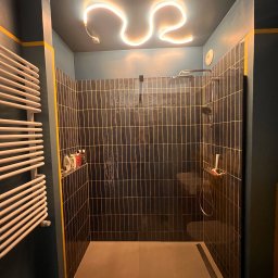 Yolo Bartosz Antonik - Nowoczesna kabina prysznicowa z czarnymi, pionowymi płytkami z żółtymi fugami, podświetlona neonowym światłem w kształcie fali na suficie, obok grzejnik drabinkowy.