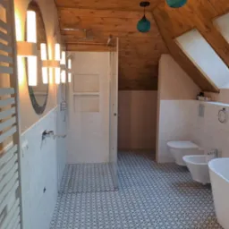 Nowoczesna łazienka na poddaszu z drewnianym sufitem, kabiną prysznicową typu walk-in, umywalką, bidetem i toaletą, oświetlona designerskimi lampami w kształcie kuli.