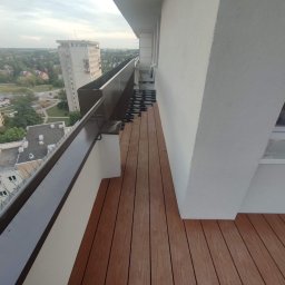 Yolo Bartosz Antonik - Montaż brązowych desek kompozytowych na balkonie, widoczne elementy mocujące i panorama miasta w tle.