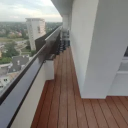 Montaż brązowych desek kompozytowych na balkonie, widoczne elementy mocujące i panorama miasta w tle.