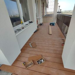 Yolo Bartosz Antonik - Montaż tarasu kompozytowego na balkonie: widoczne deski, narzędzia (wkrętarka, miarka, ołówek), skrzynki z łącznikami, częściowo ułożona podłoga, widok na miasto w tle.