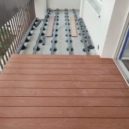 Yolo Bartosz Antonik - Montaż brązowego tarasu kompozytowego na balkonie, widoczne legary i podkładki poziomujące, fragment balustrady i zabezpieczone folią okno.