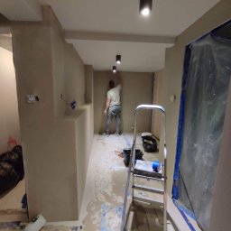 Yolo Bartosz Antonik - Mężczyzna w trakcie szpachlowania ściany w korytarzu, widoczne przygotowane podłoże, drabina i zabezpieczone folią drzwi.