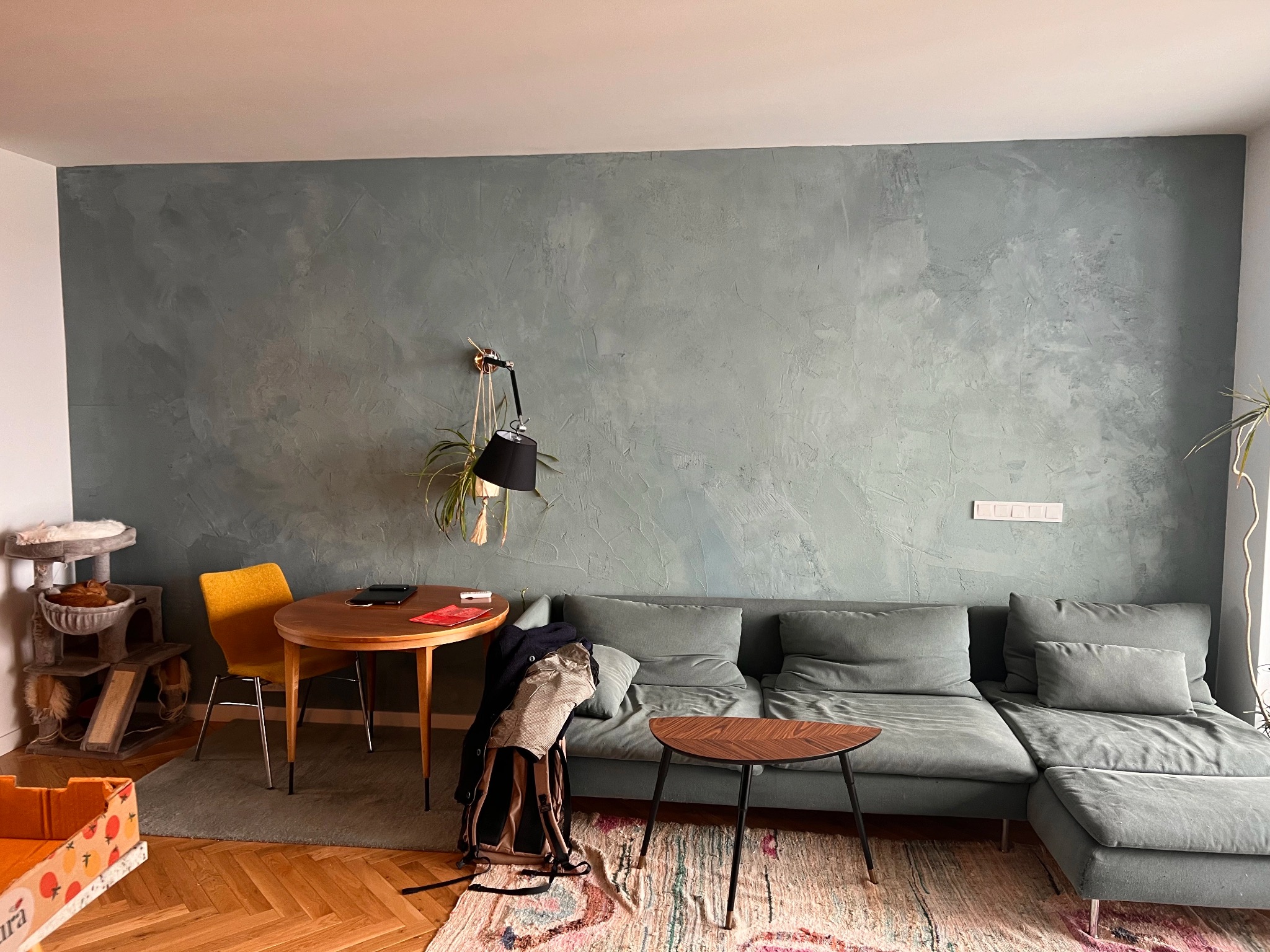 Ściana w odcieniach szarości z dekoracyjnym efektem malarskim, uzupełniona o minimalistyczną lampę ścienną, stolik kawowy, szarą sofę i dywan w pastelowych kolorach.