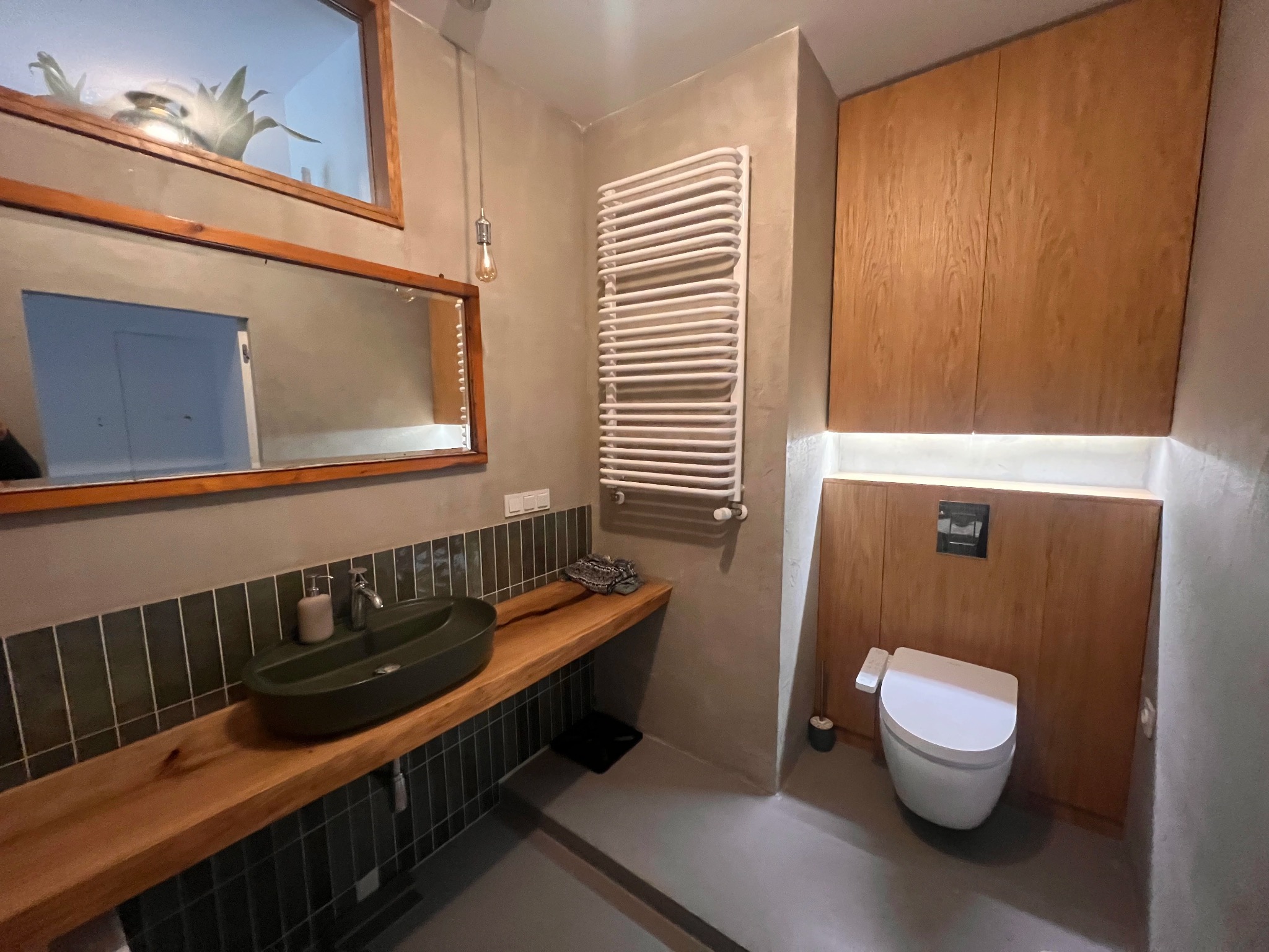 Nowoczesna łazienka z drewnianym blatem pod umywalką w kolorze oliwkowym, grzejnikiem drabinkowym i toaletą w zabudowie z drewnianymi frontami.