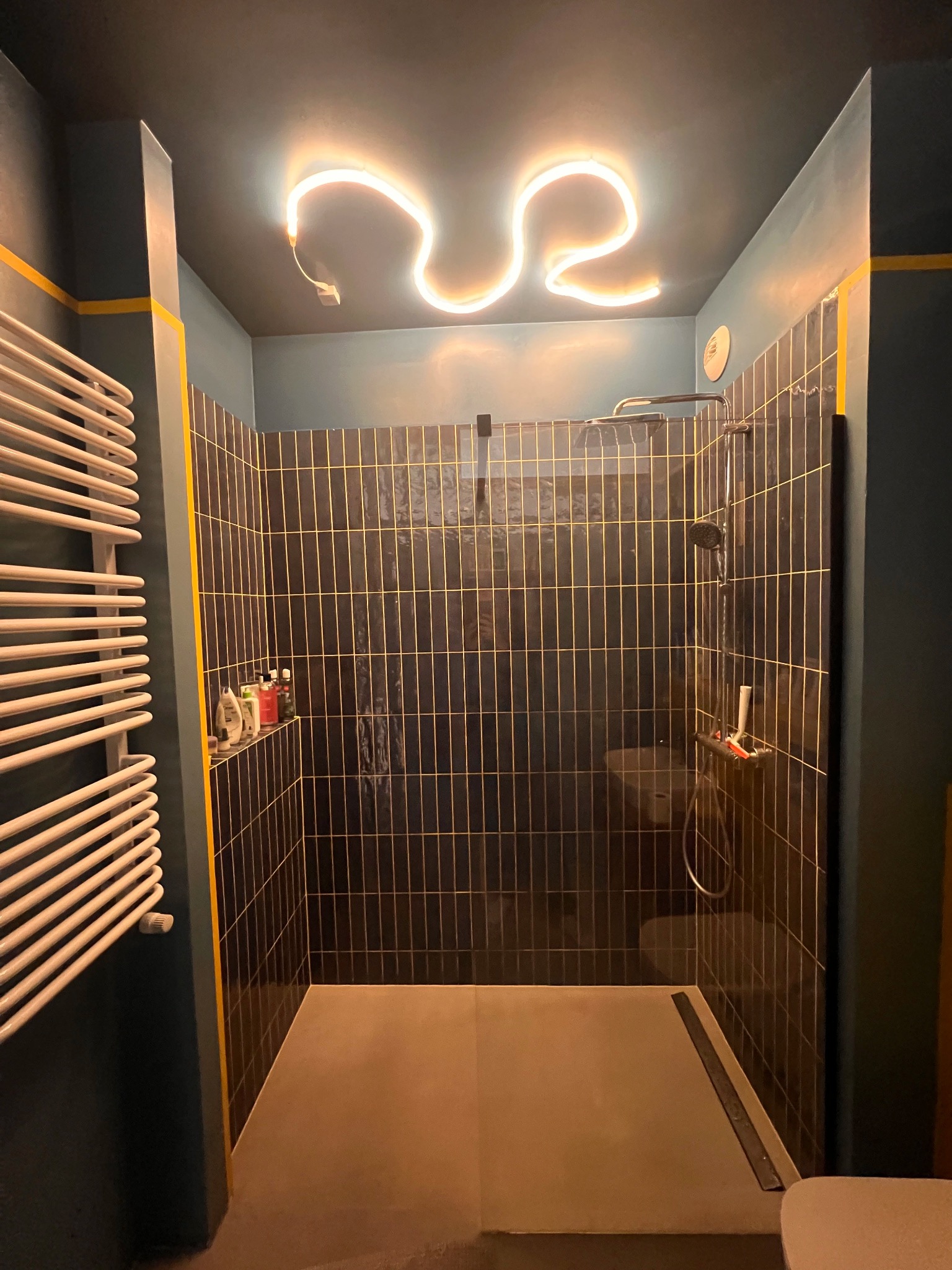 Nowoczesna kabina prysznicowa z czarnymi, pionowymi płytkami z żółtymi fugami, podświetlona neonowym światłem w kształcie fali na suficie, obok grzejnik drabinkowy.