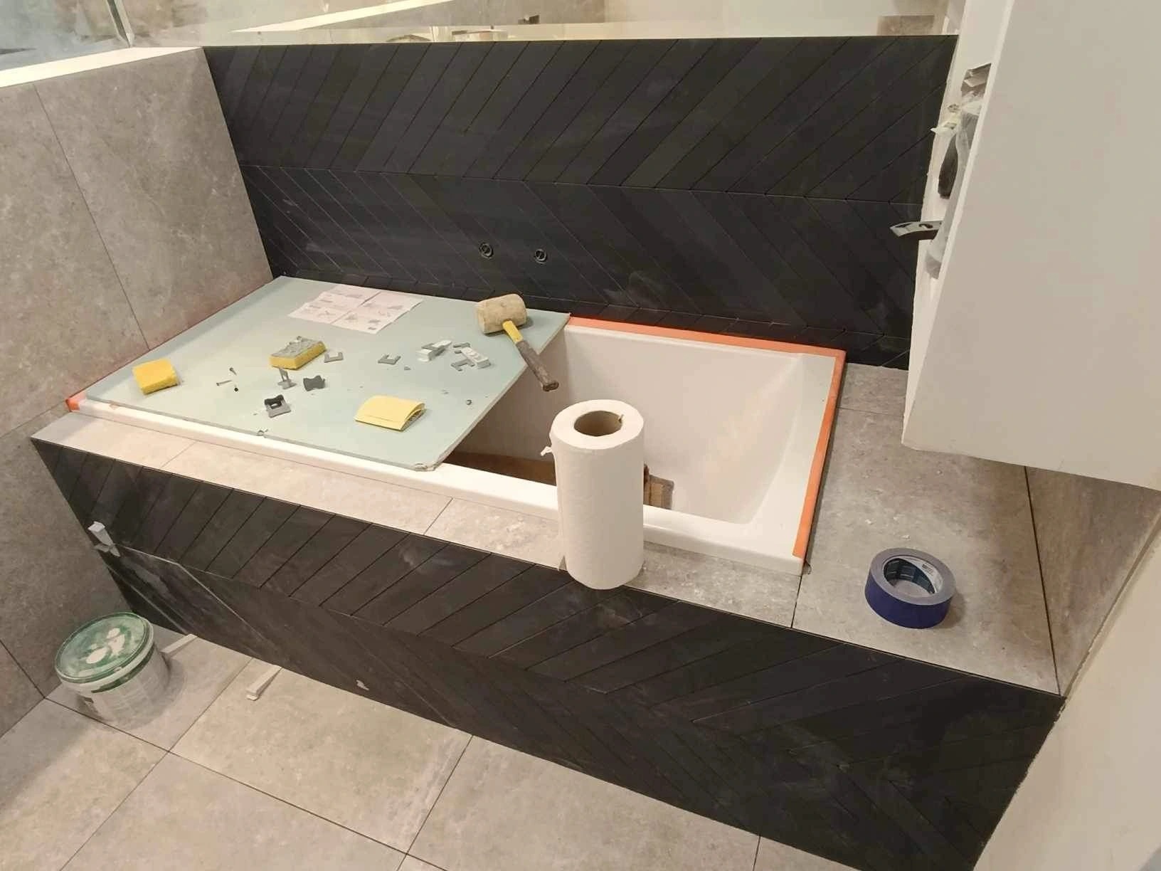 Montaż płytek chevron w obudowie wanny, widoczne narzędzia glazurnicze, poziomica i rolka papieru.