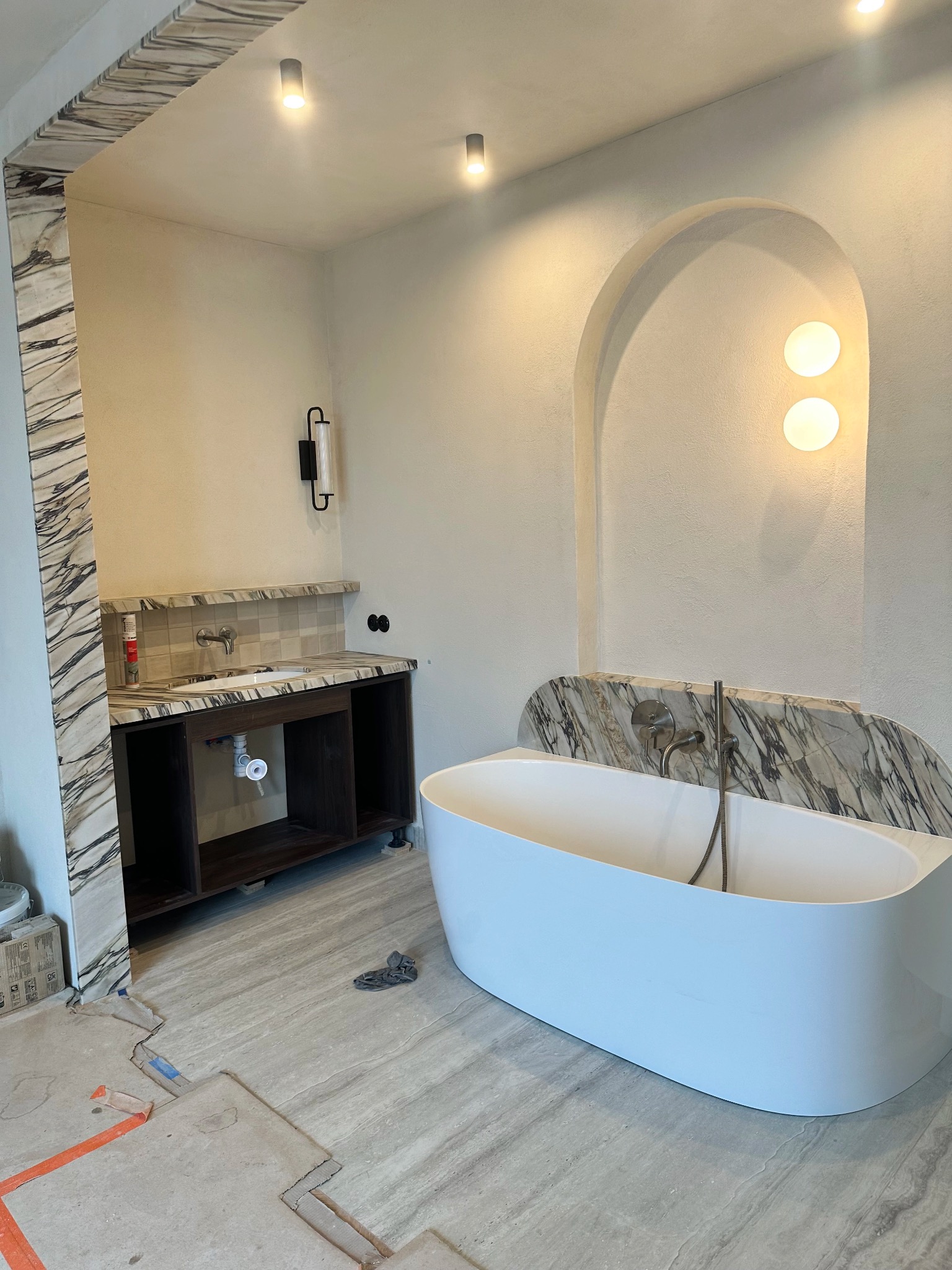 Nowoczesna łazienka w trakcie wykańczania: biała wanna wolnostojąca z marmurowym panelem, szafka pod umywalkę z ciemnego drewna i marmurowym blatem, podłoga wyłożona płytkami imitującymi marmur...