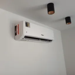 Biała jednostka klimatyzacji Whirlpool zamontowana na białej ścianie, widoczne dwa czarne reflektory sufitowe z miedzianym wnętrzem.