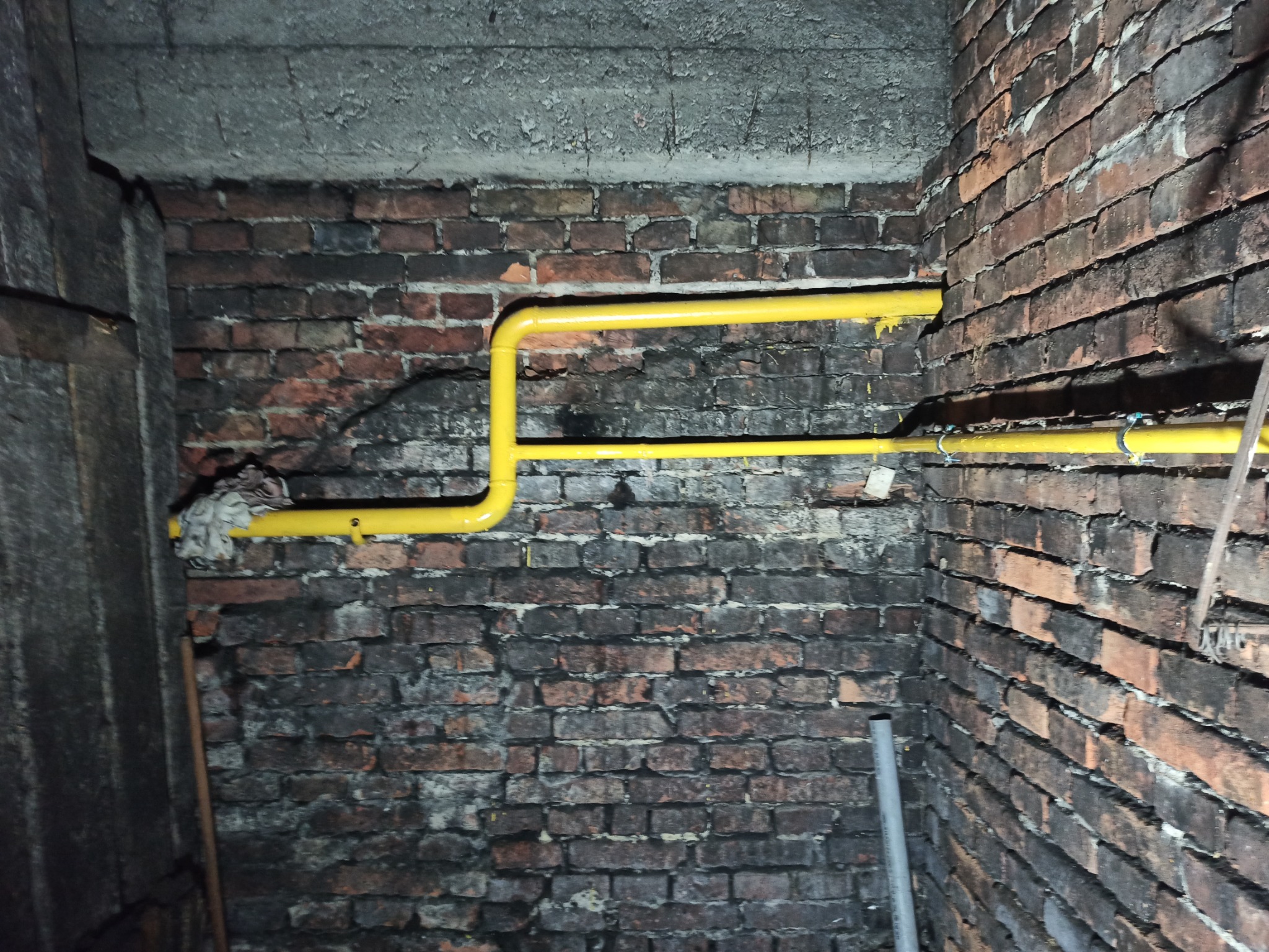 Żółte rury wentylacyjne zamontowane na starej, ceglanej ścianie w piwnicy, widoczne mocowania i fragment szmaty.