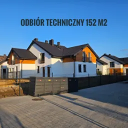 Widok na nowo wybudowane domy jednorodzinne z drewnianymi elementami elewacji, widoczne pojemniki na odpady i kostka brukowa na podjeździe, w tle błękitne niebo.