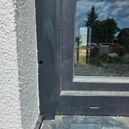Usługi Inżynierskie mgr inż. Monika Wójcicka - Szczegółowe ujęcie osadzonego okna PCV w szorstkiej, jasnej elewacji budynku z widocznym otworem montażowym i refleksami otoczenia na szybie.