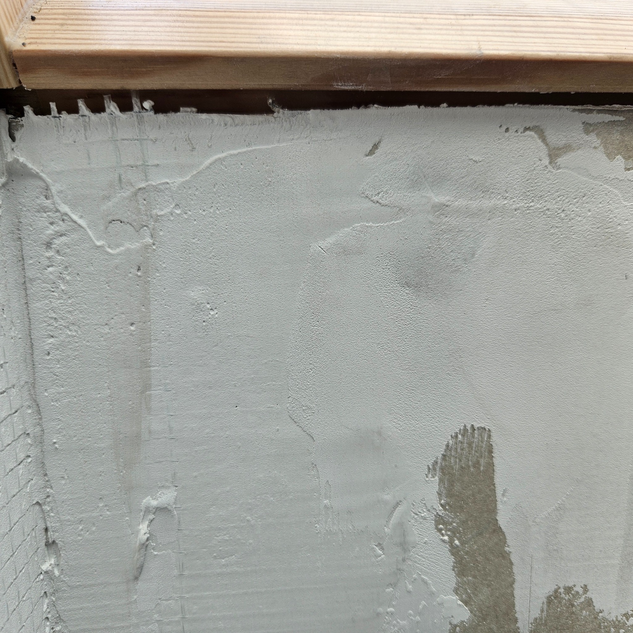 Świeżo nałożona, szara warstwa tynku na ścianie z widoczną siatką wzmacniającą w lewym dolnym rogu, drewniana listwa u góry.
