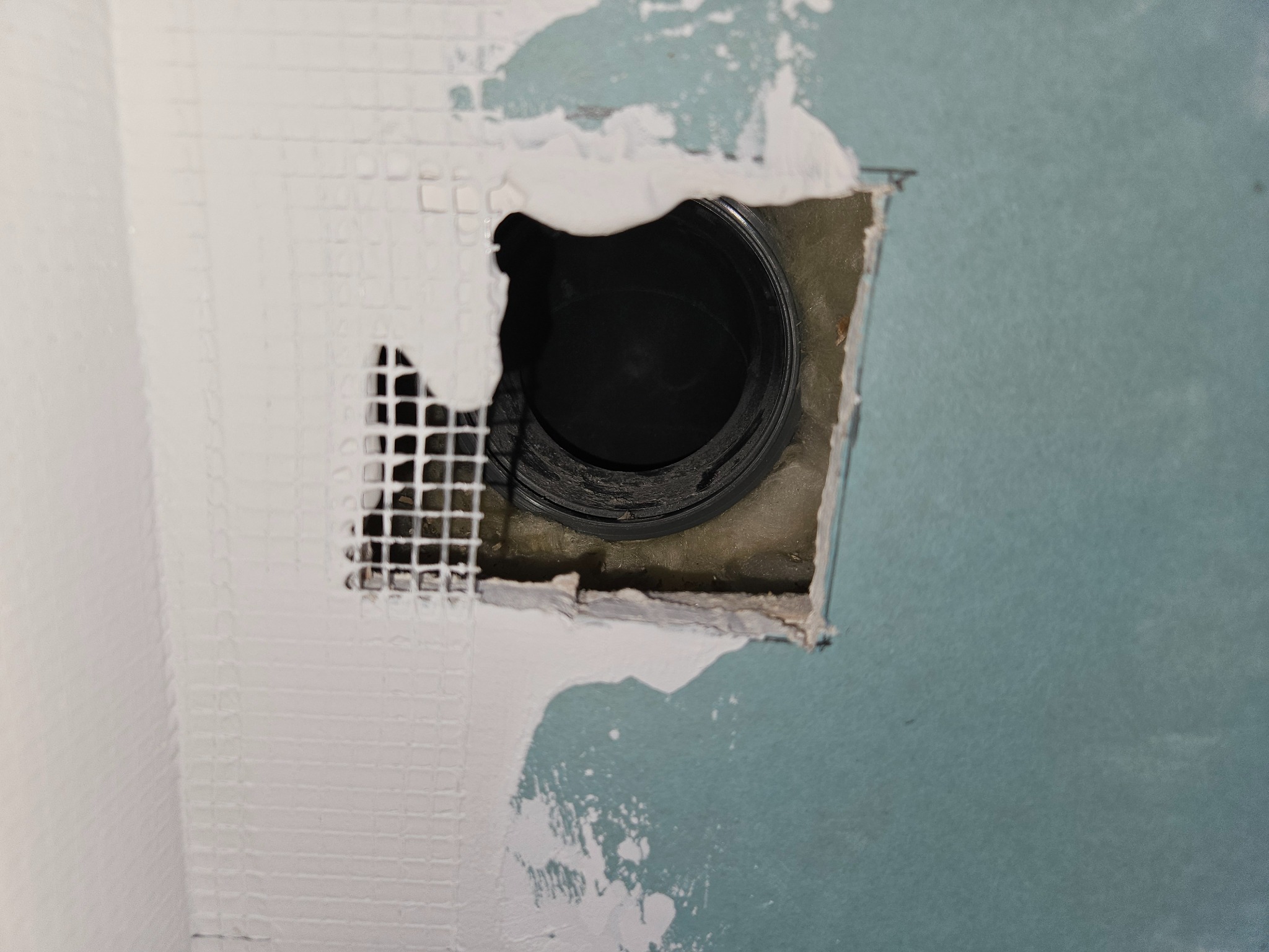 Otwór w ścianie z widoczną czarną rurą wentylacyjną, częściowo zakryty siatką wzmacniającą i gipsem, ściana pomalowana na jasnoniebiesko.