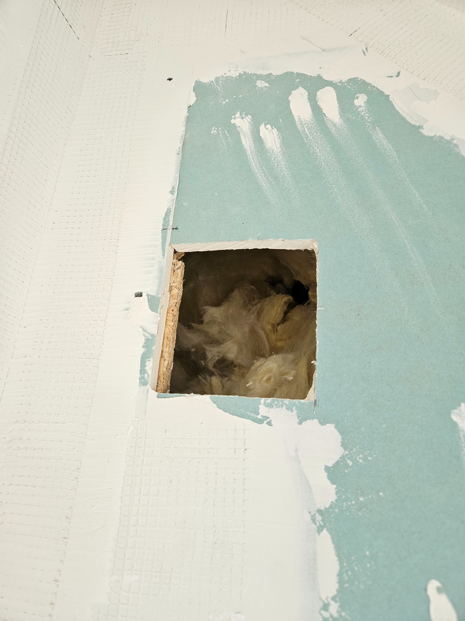 Otwór w płycie gipsowo-kartonowej, odsłaniający izolację z wełny mineralnej. Widoczne ślady szpachlowania na sąsiednich płytach.