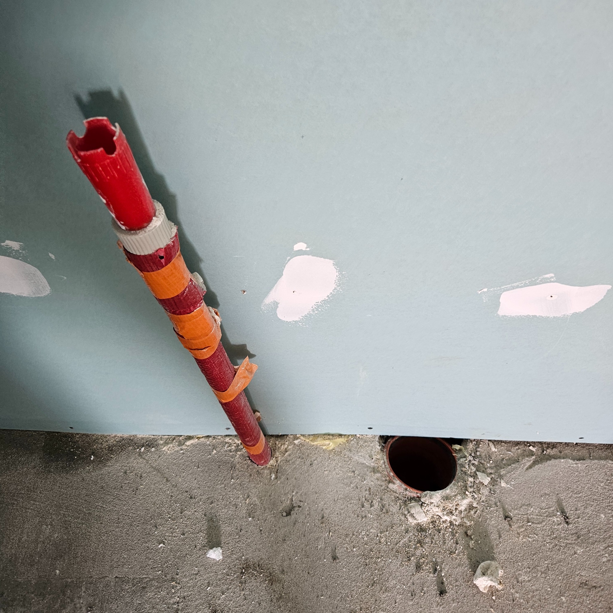 Czerwona rura instalacyjna z pomarańczową taśmą, wystająca ze ściany z płyt gipsowo-kartonowych, widoczne ślady szpachlowania i otwór w podłodze.